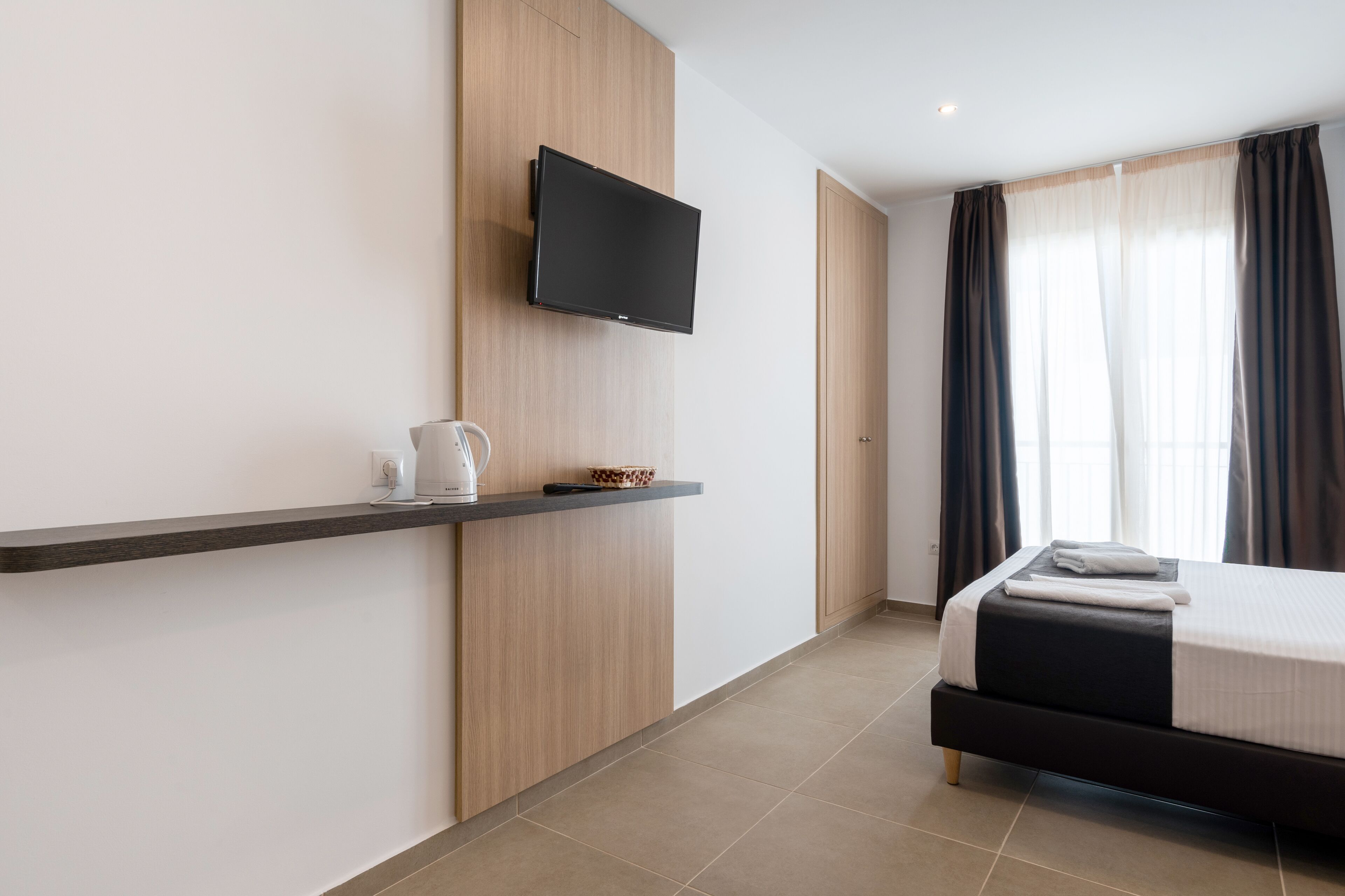 Una habitación de hotel moderna con una cama, un televisor montado, una cafetera y un estante con una tetera y un cuenco decorativo.