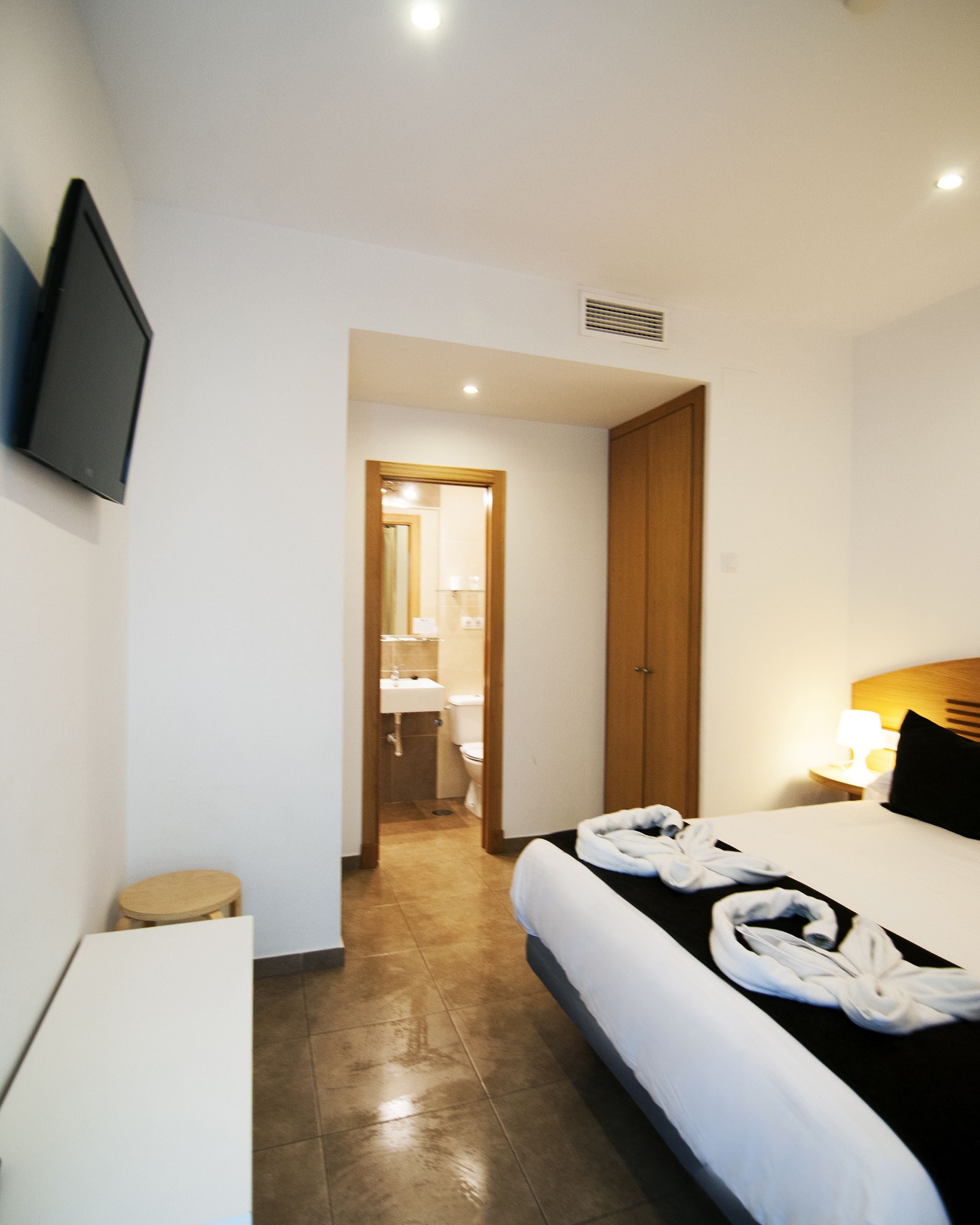 Habitación de hotel con cama, escritorio, silla, televisor montado en la pared y baño visible a través de una puerta abierta.