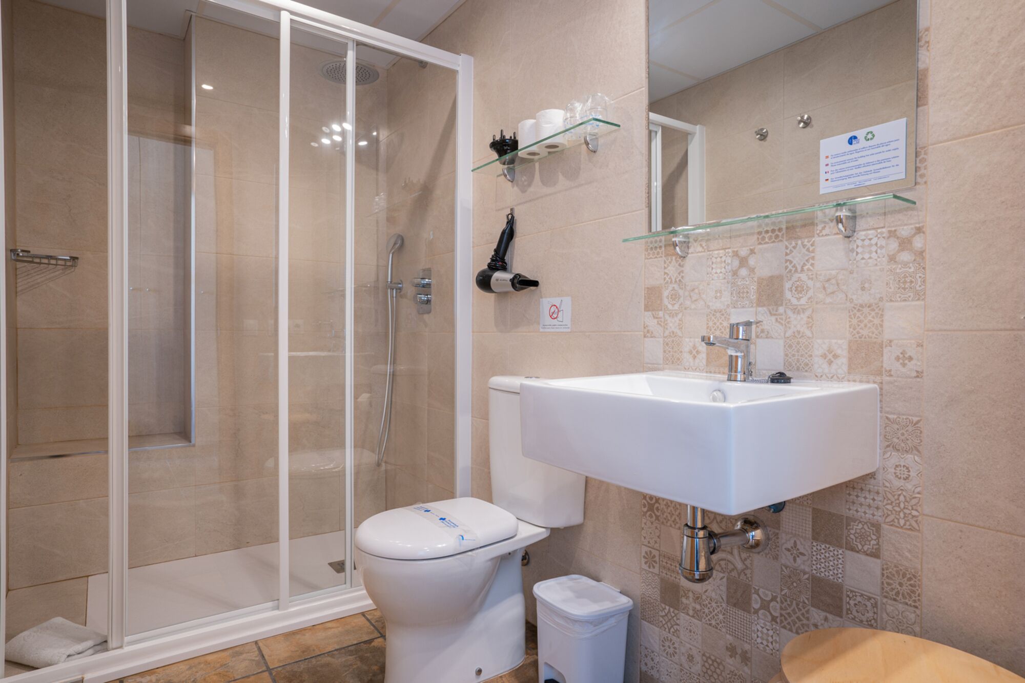 Une salle de bain dotée d’une douche à parois de verre, de toilettes blanches, d’un lavabo avec un miroir au-dessus et de carreaux décoratifs sur les murs.