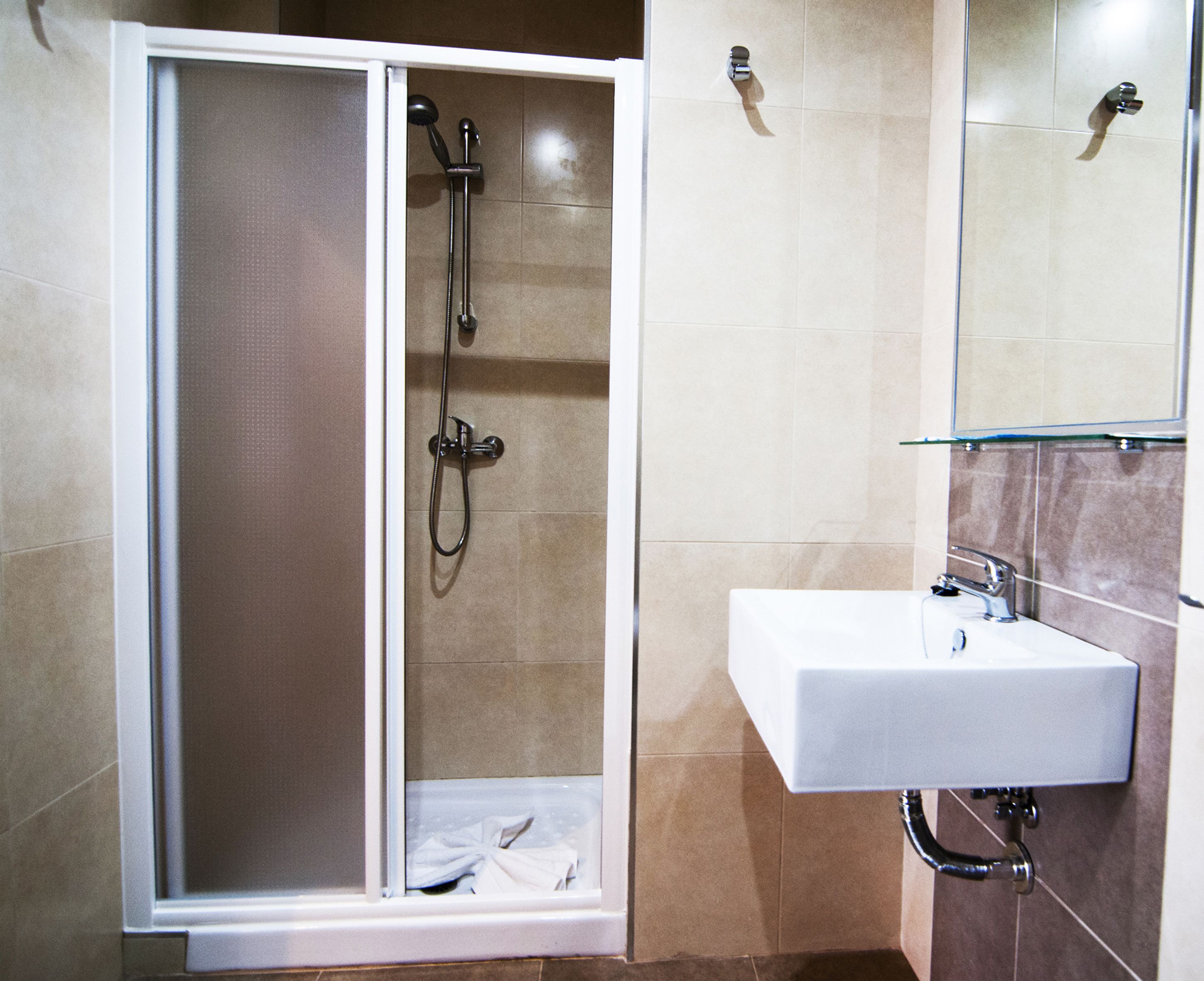 Un espace douche doté d’une porte en verre, d’un lavabo blanc avec un miroir au-dessus, et de murs recouverts de carreaux beiges.