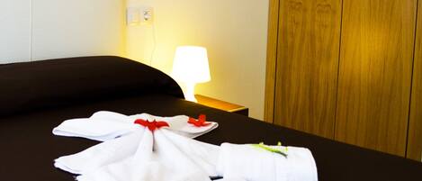 Siististi pedattu sänky, jossa pyyhkeet on aseteltu viuhkamaisesti, yöpöydällä oleva lamppu ja puinen vaatekaappi hotellihuoneessa.
