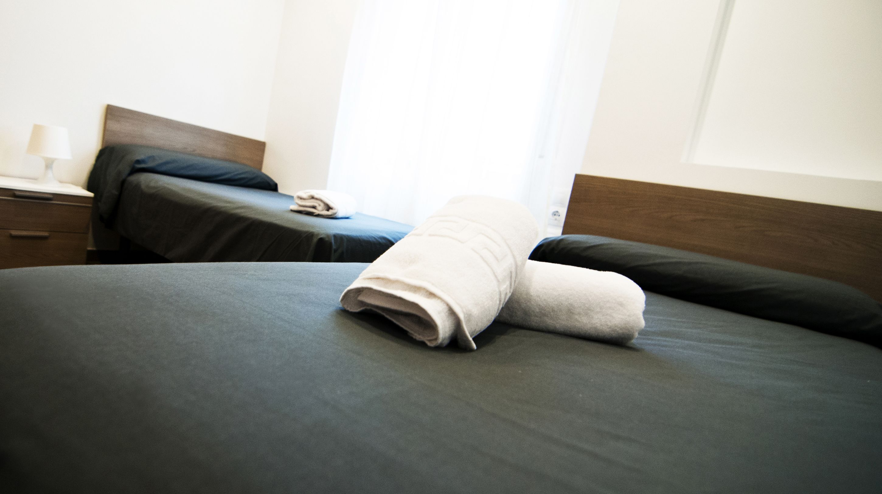 A neatly made bed with a folded towel, a bedside table with a lamp, and a nightstand with a phone.