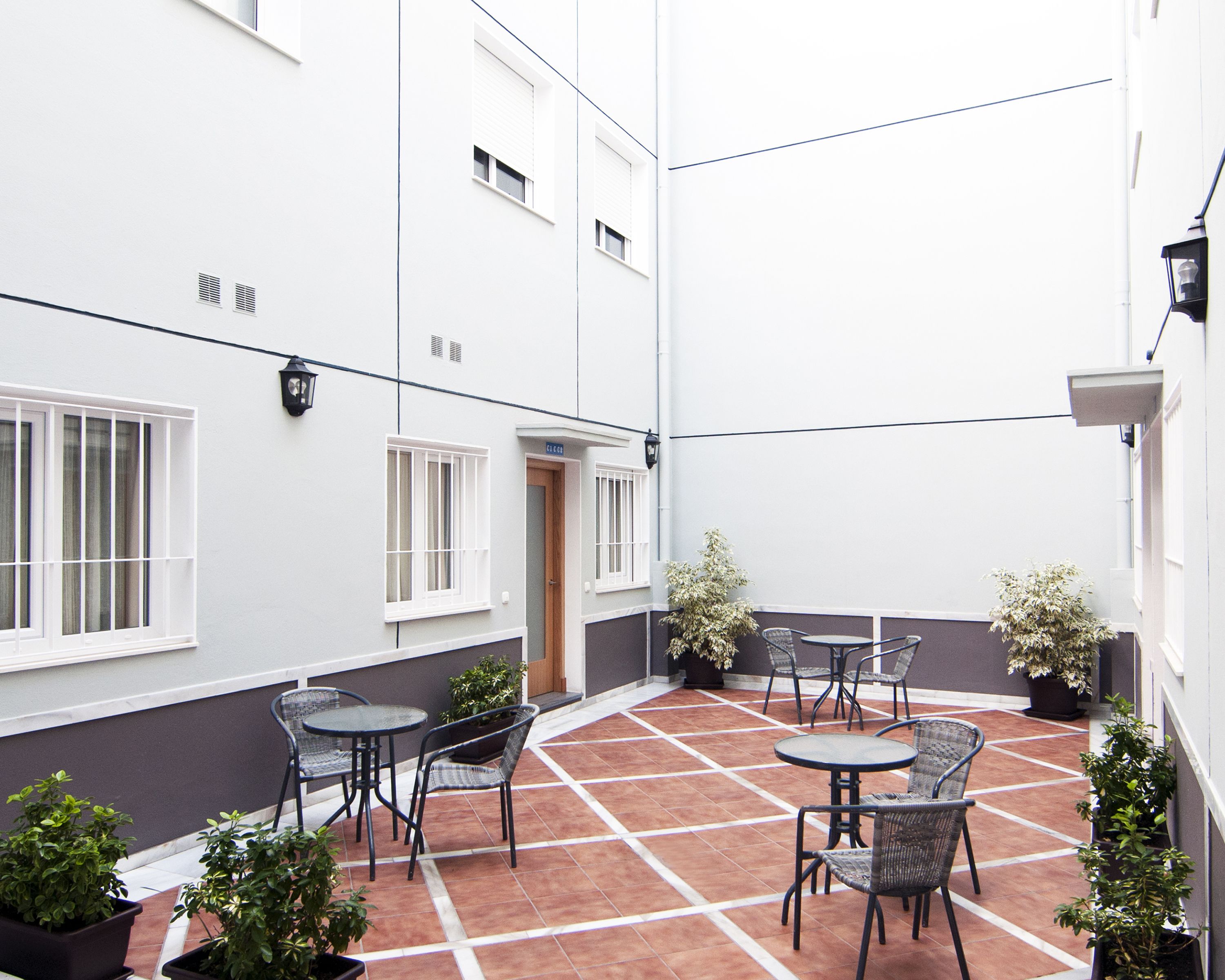 A courtyard with tiled flooring, outdoor seating, potted plants, and a building with white walls and windows.