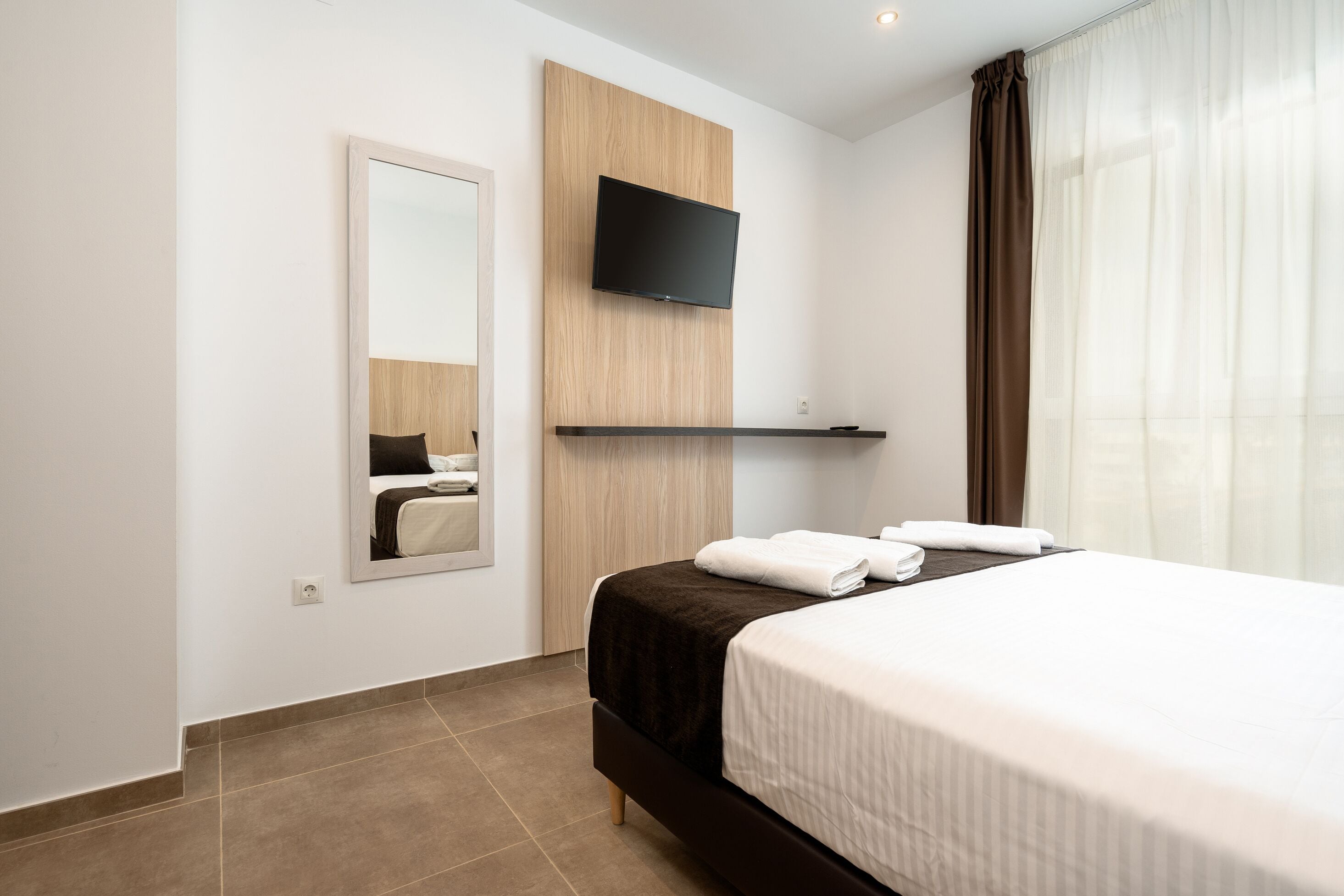 A hotel room with a bed, a mounted TV, a mirror, and a window with curtains.
