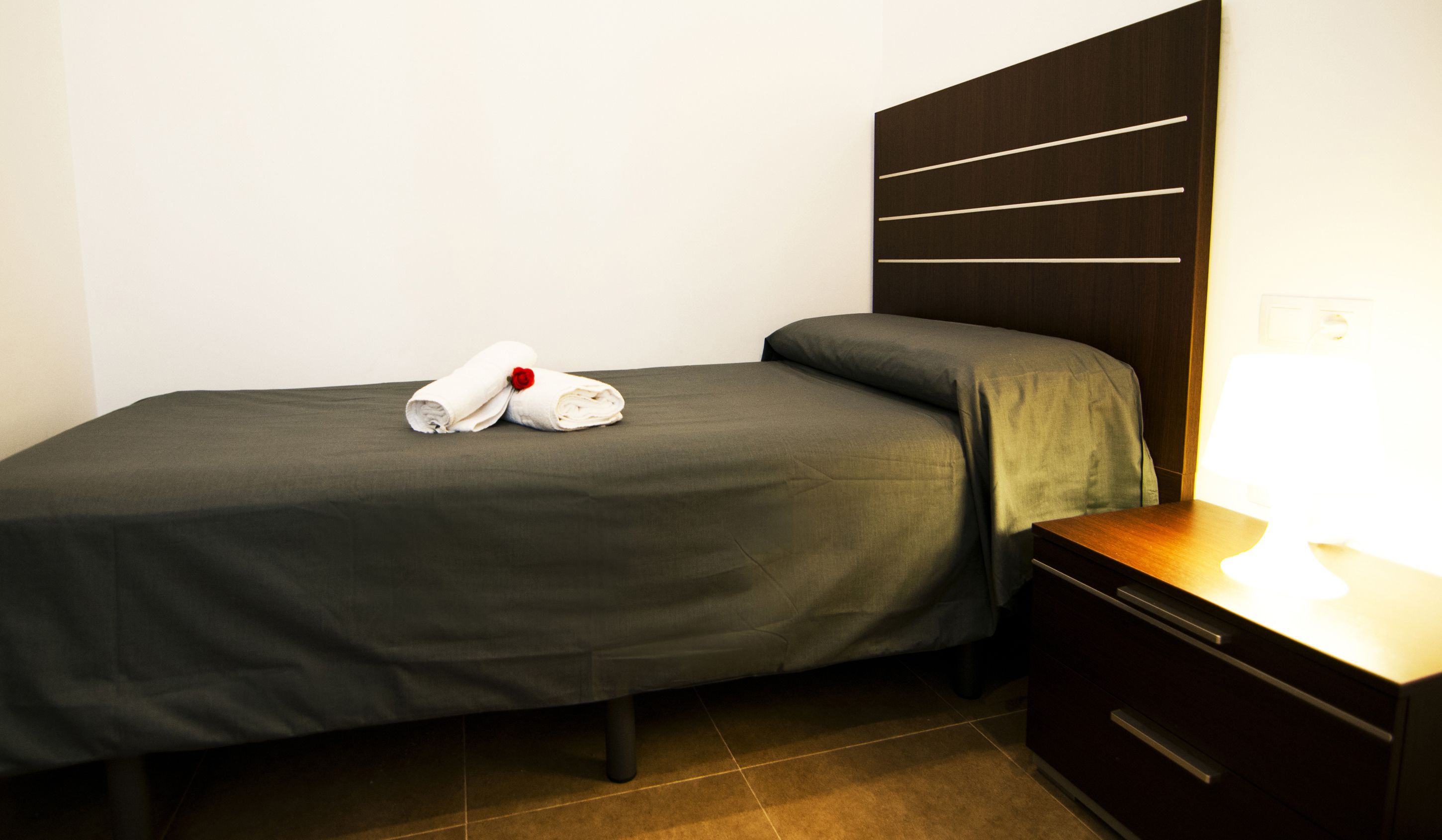 Ein ordentlich bezogenes Bett mit einer grauen Tagesdecke und Kissen, ein Nachttisch aus Holz mit einer Lampe und ein dunkles, horizontal-liniertes Holz-Kopfteil.