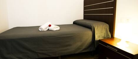 Una cama bien tendida con colcha y almohadas grises, una mesita de noche de madera con una lámpara, y un cabecero de madera oscura con líneas horizontales.