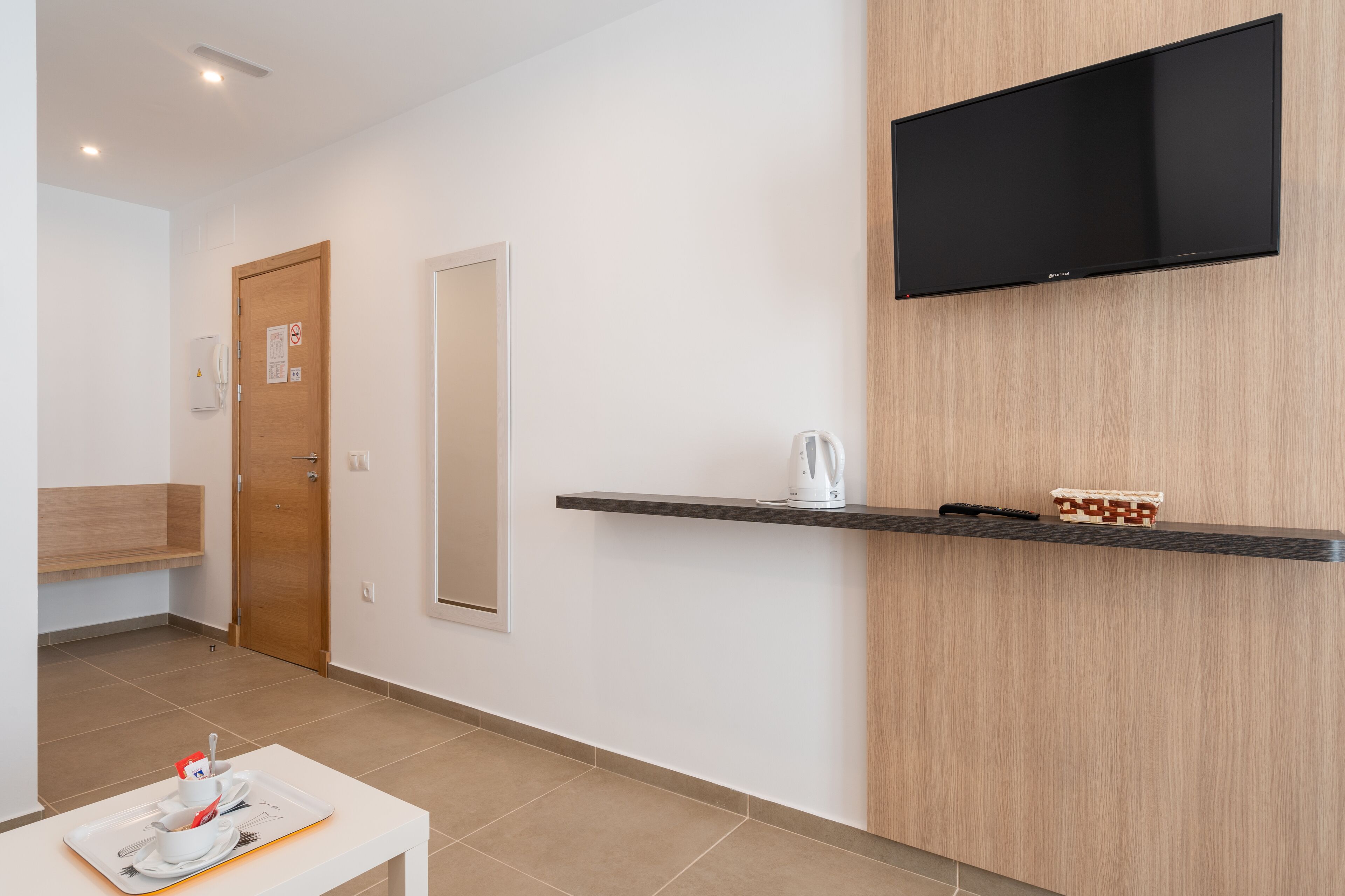 Una habitación de hotel moderna con una puerta de madera, un televisor de pantalla plana montado en la pared, un estante con un hervidor y un objeto decorativo, y una bandeja con una taza y un plato sobre una mesa.