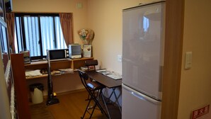 A compact room with a wooden table, two chairs, a small desk with a computer and a microwave, a refrigerator, and a window with curtains. - Daiya Ryokan (Kyoto)