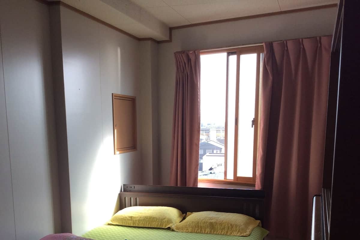 A hotel room with a bed, a desk, a chair, a window with curtains, and a view of buildings outside.