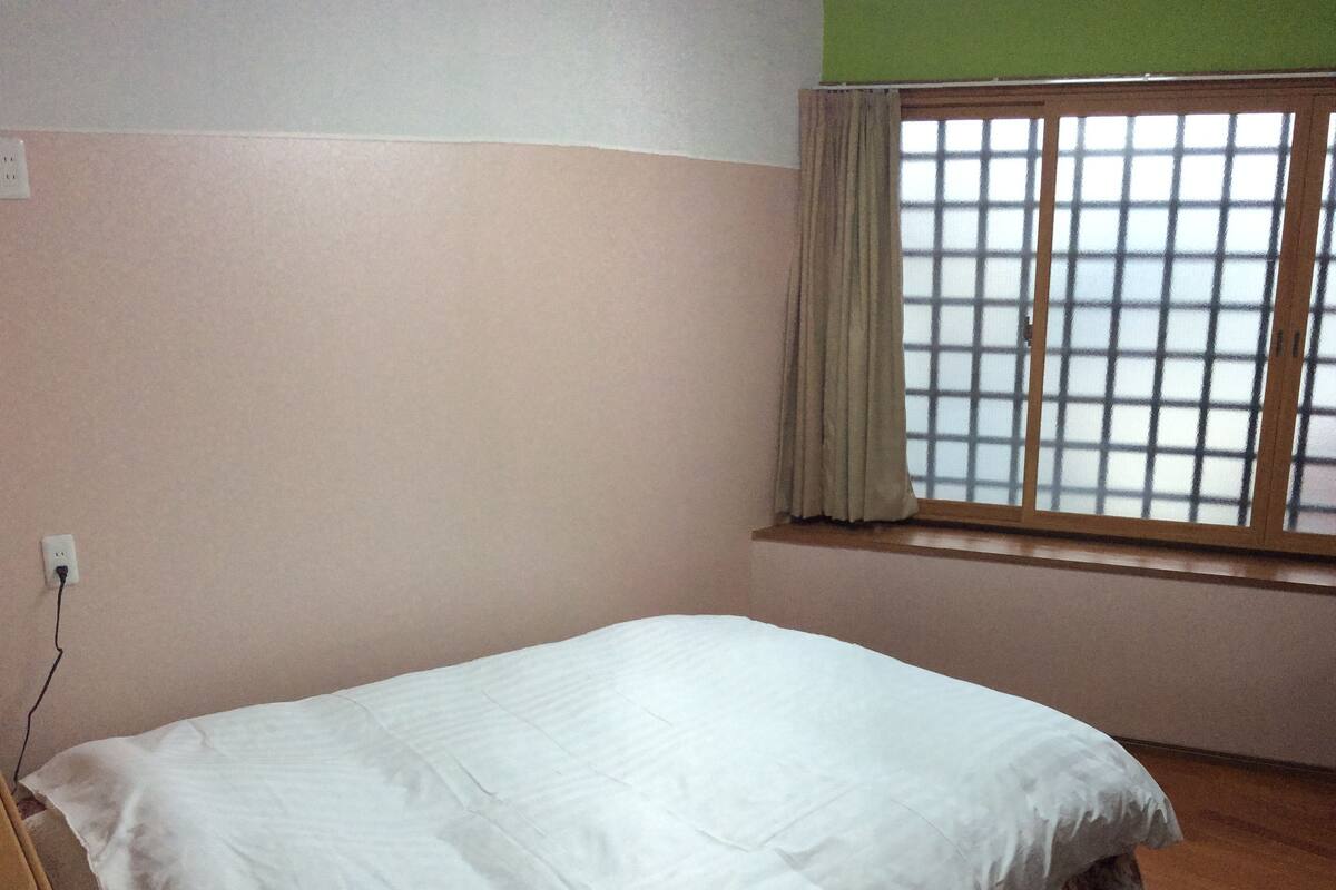 A room with a single bed, a window with a grid pattern, and a power outlet on the wall.