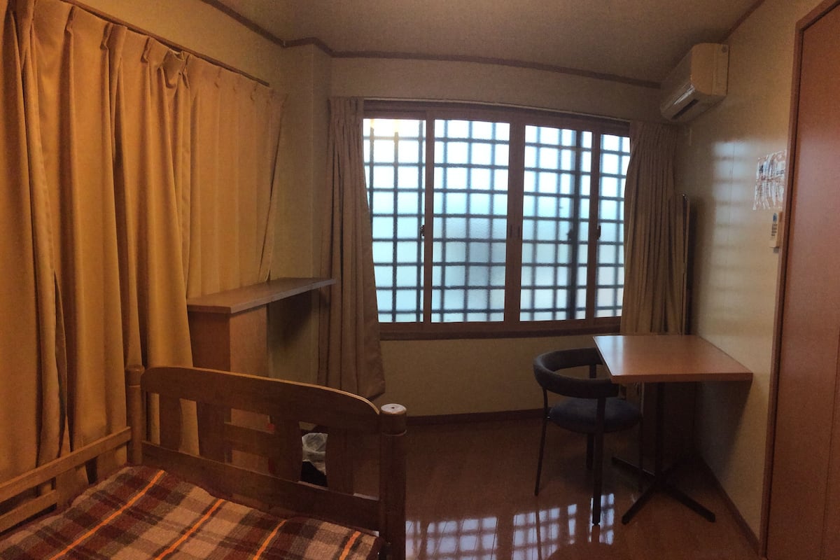 A small room with a bed, a desk, a chair, and a window with curtains.