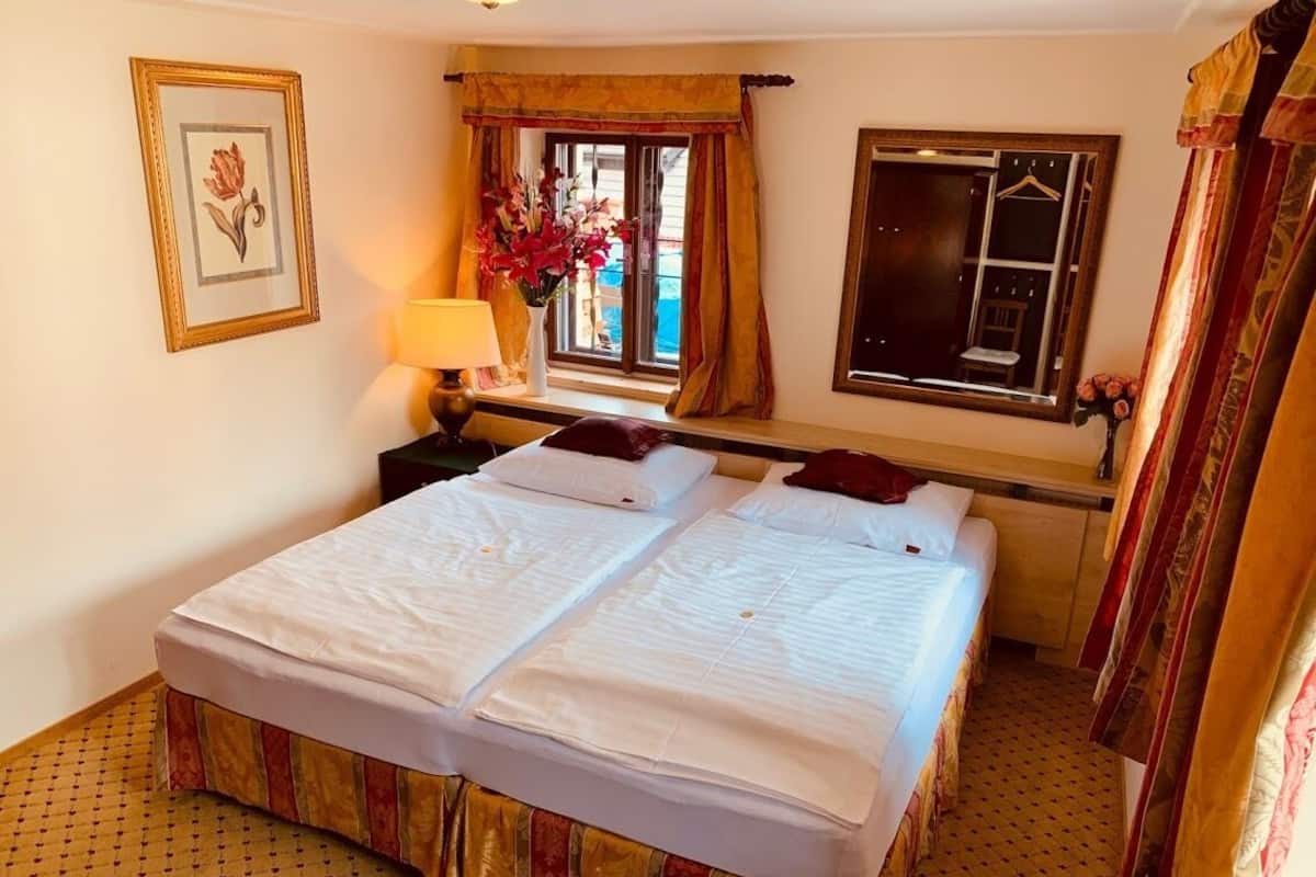 A double bed with white linens, a nightstand with a lamp, a window with curtains, and a framed picture on the wall.