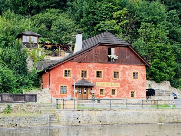 A red building with a sign reading "PENZION KAPR" near a body of water, surrounded by greenery. - Penzion Kapr (Cesky Krumlov)