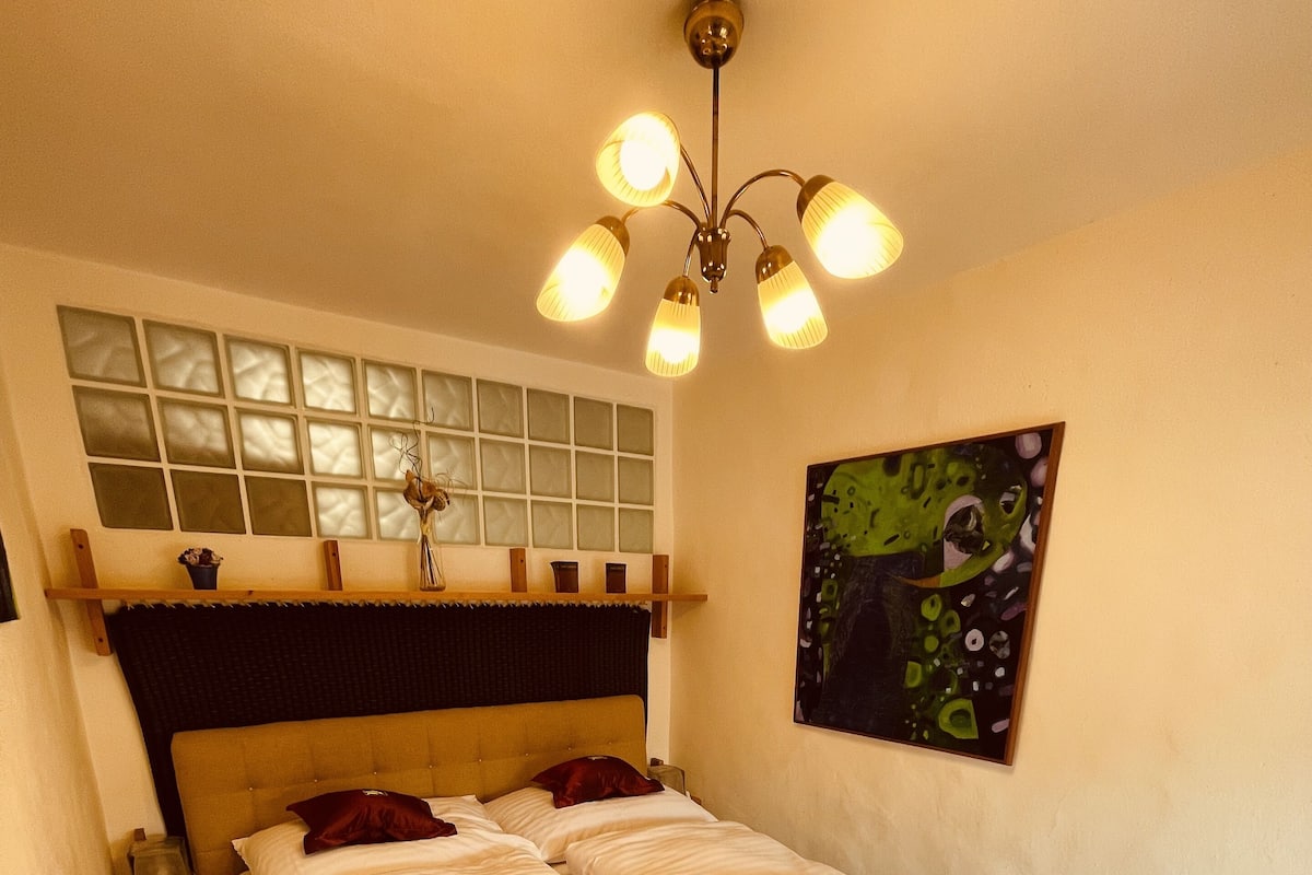 A hotel room with a large bed, a wooden headboard, a ceiling light fixture, a window with blinds, and a painting on the wall.
