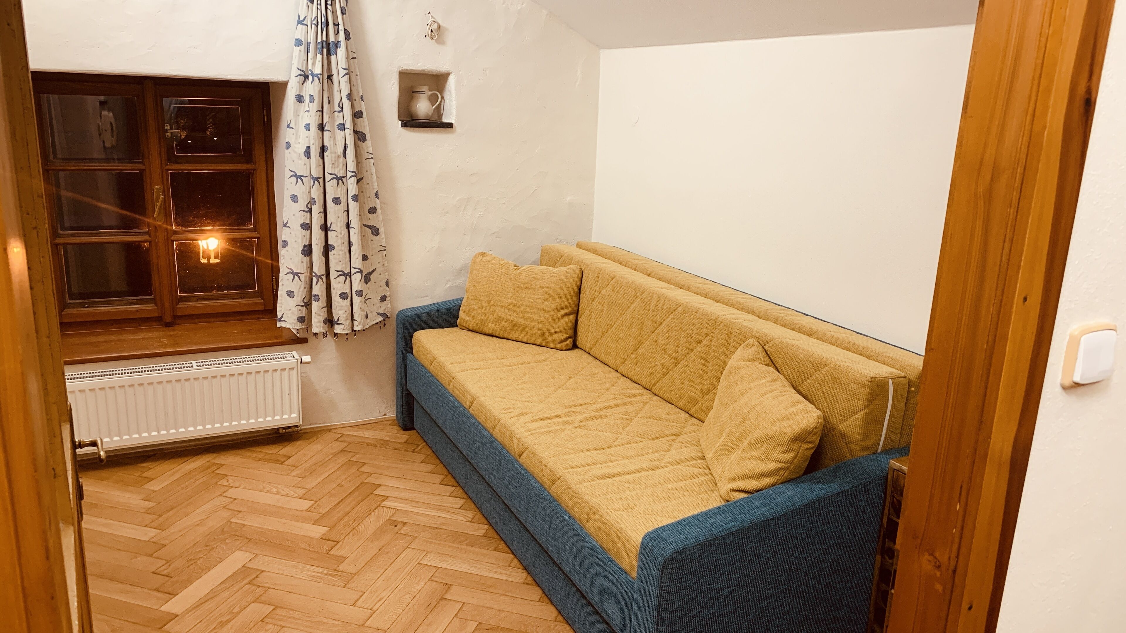 Une pièce chaleureuse avec un plancher en bois, un canapé beige orné de bordures bleues, une fenêtre avec des rideaux et un radiateur blanc.