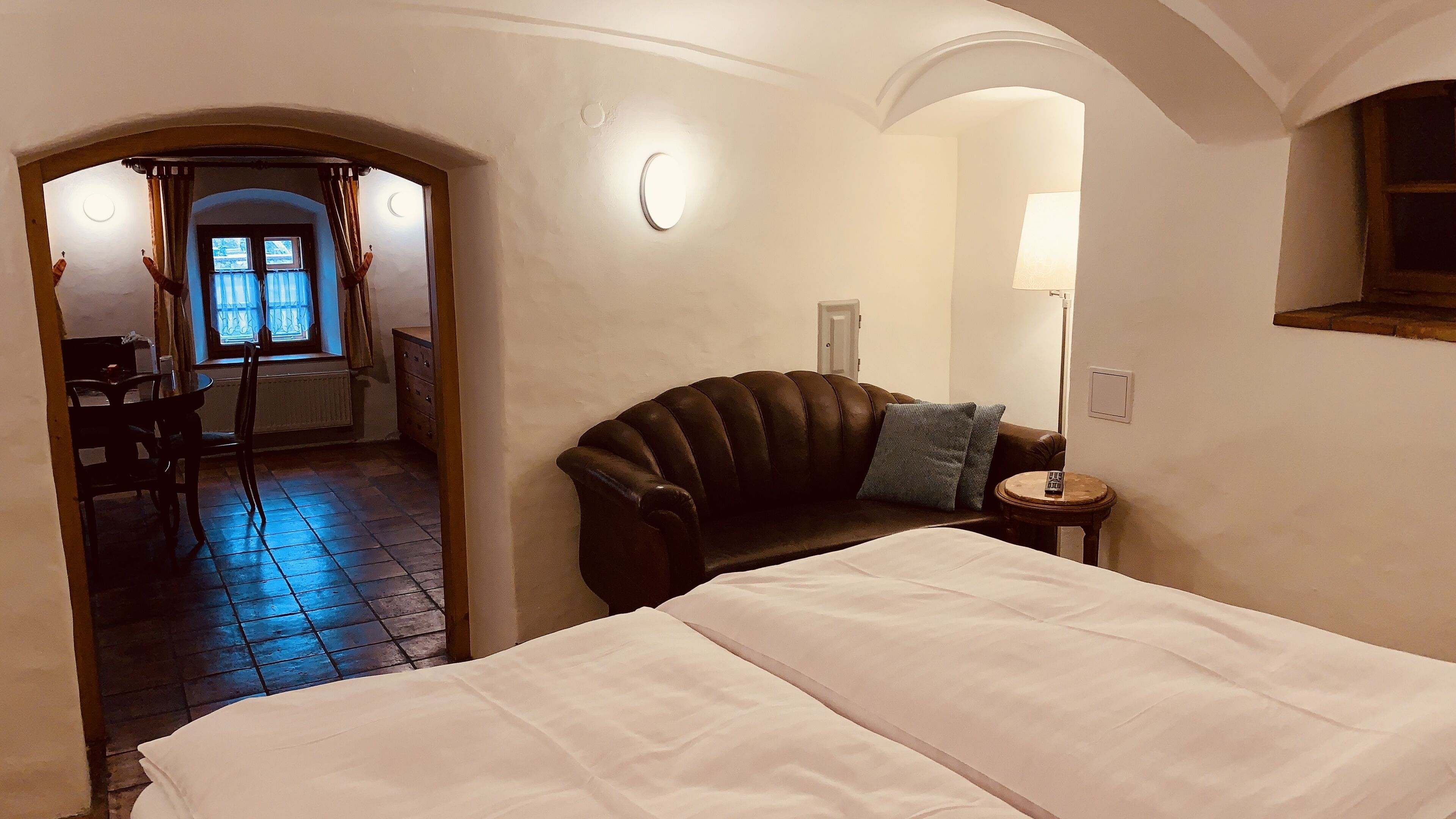 A hotel room with a bed, a leather armchair, a small table, and a lamp. There is a window with a view of the outside.