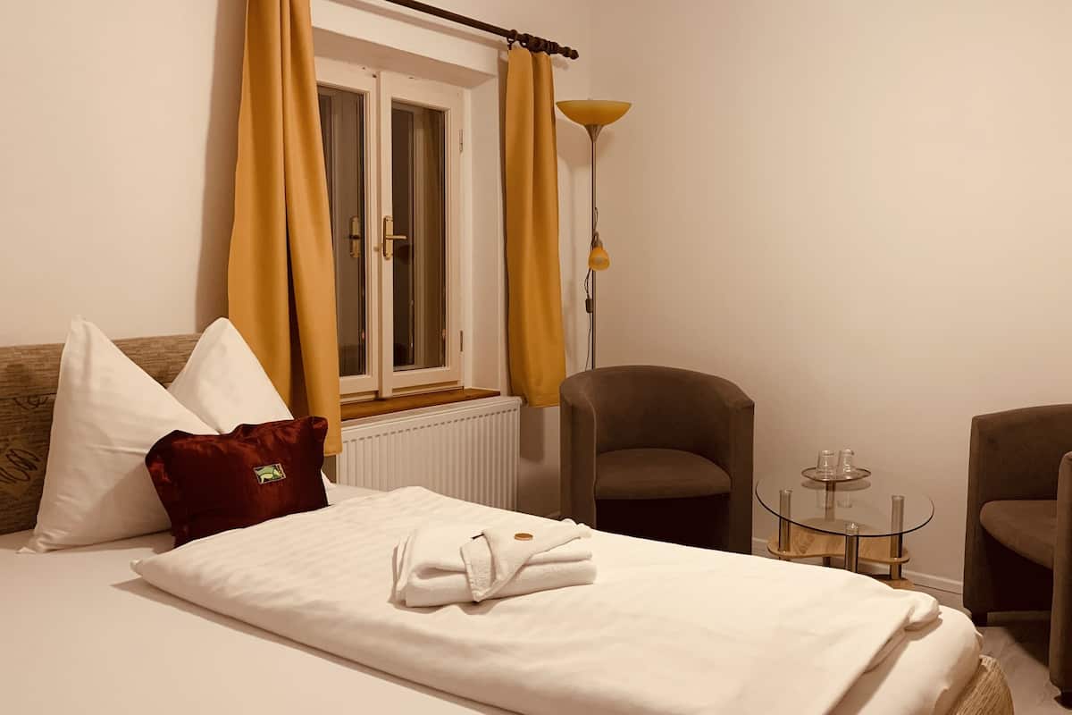 A neatly made bed with white linens and a folded towel, a brown armchair, a round table with a glass top, and a floor lamp.