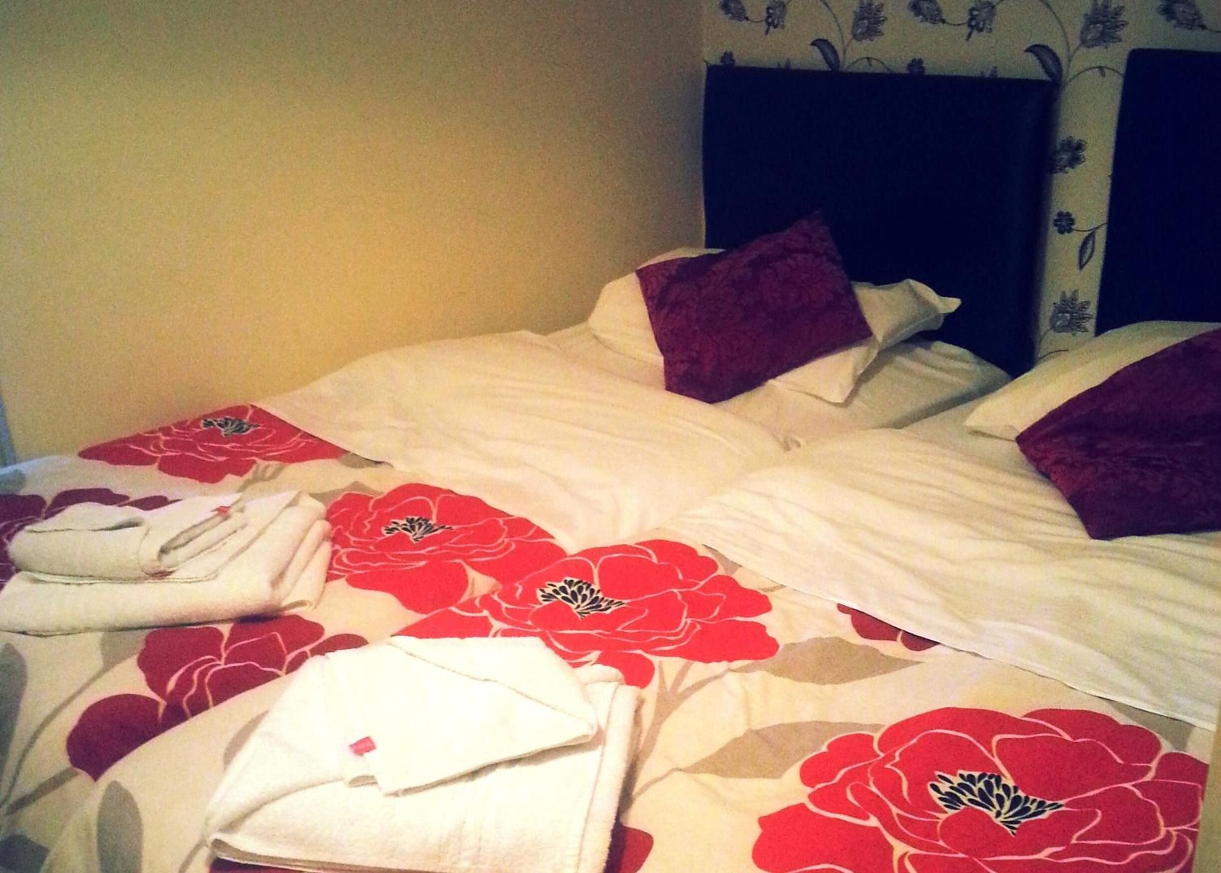 A double bed with white bedding and red floral patterns, a dark headboard, and a folded white towel on the bed.