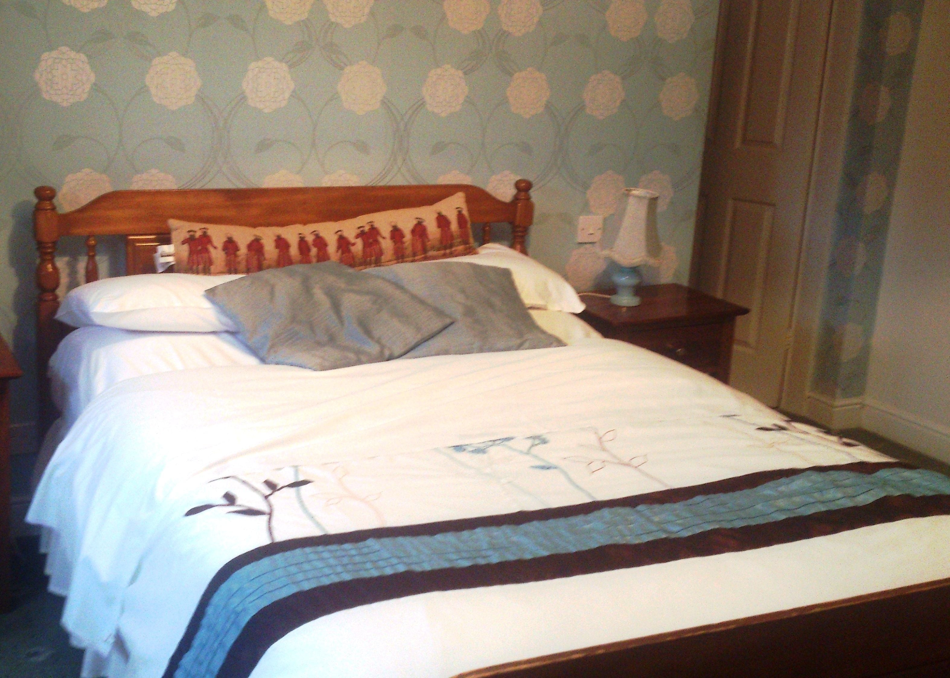 A neatly made bed with a floral-patterned headboard, a bedside table with a lamp, and a wall with a floral wallpaper design.