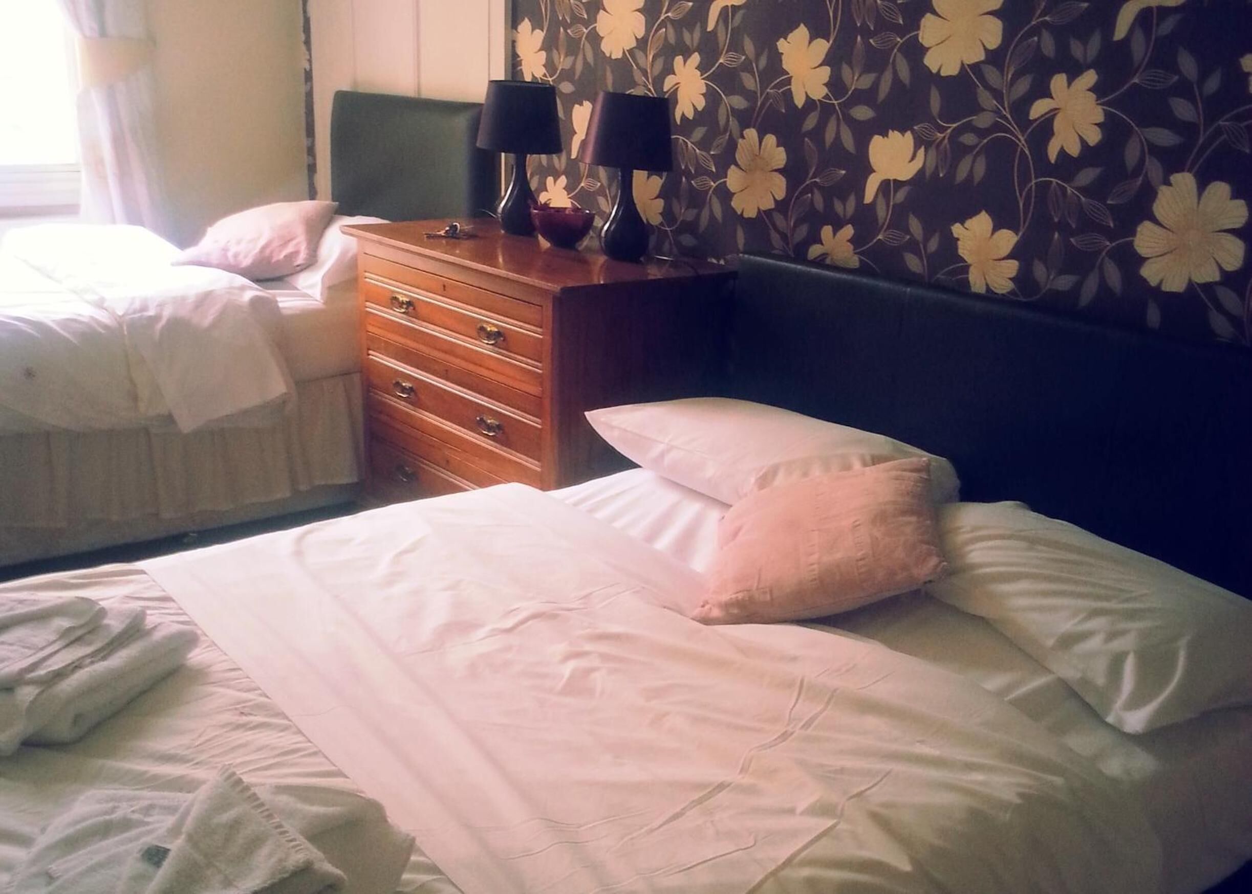 A hotel room with two beds, a wooden dresser, and a floral wallpaper.