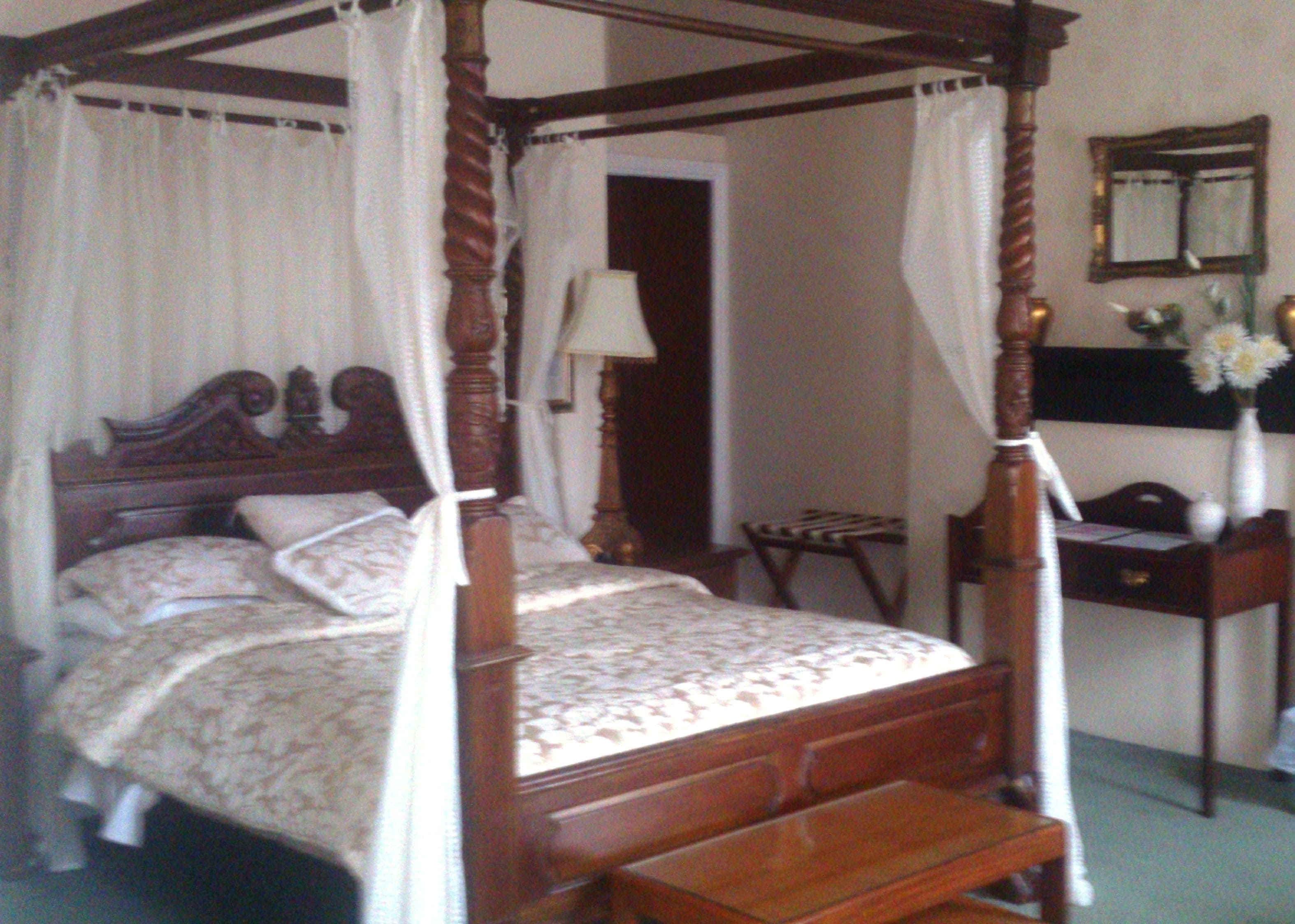 Una cama con dosel y cuatro columnas, con cabecera y piecera de madera, una mesita de noche y una silla.