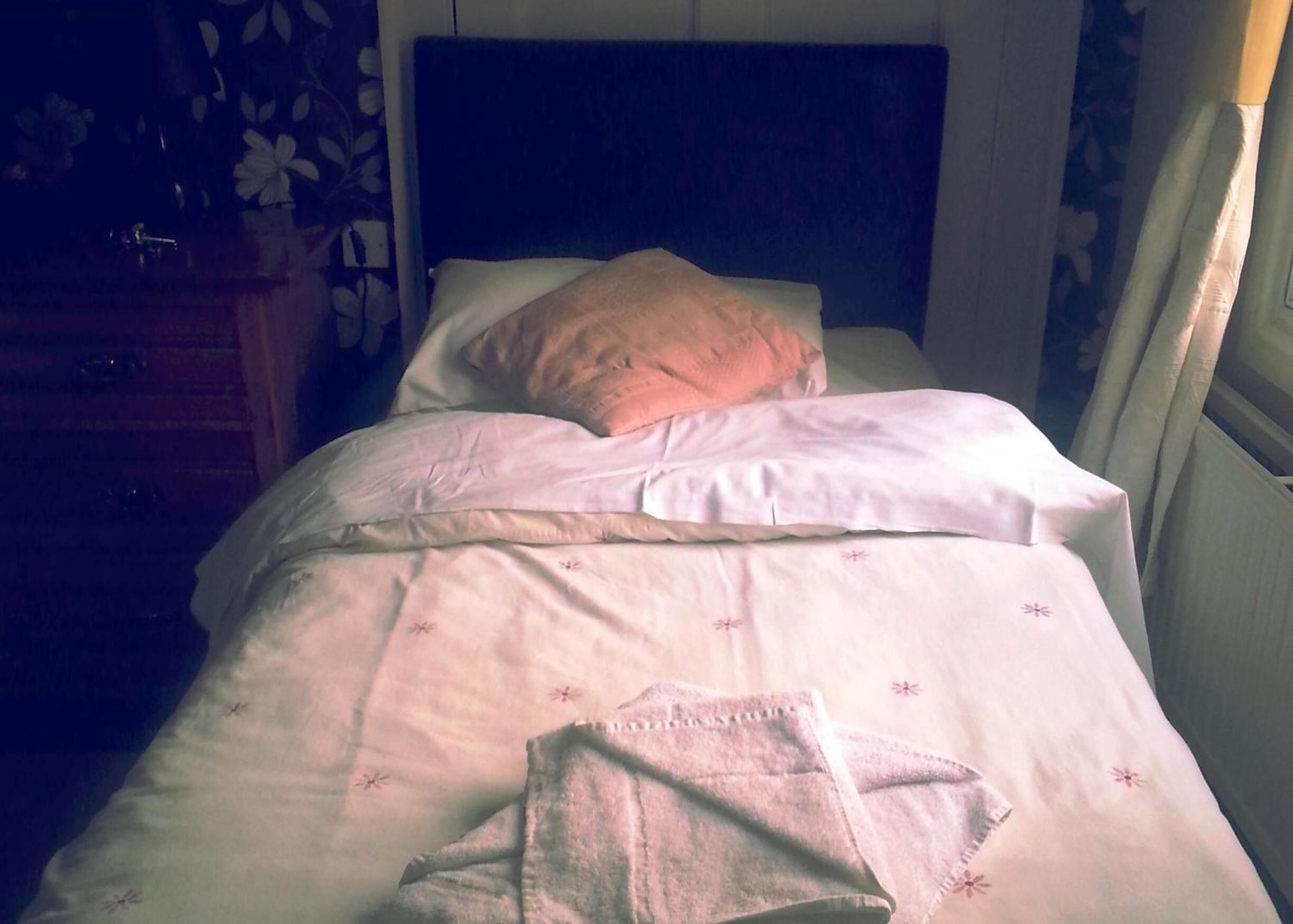 A bed with white bedding and a pillow, a wooden nightstand, and a wall with a floral pattern.