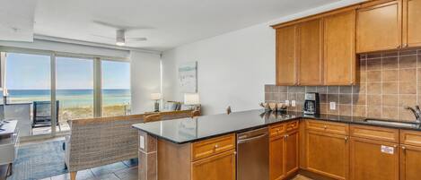 A modern kitchen with wooden cabinets, a black countertop, and a view of the ocean through large windows.