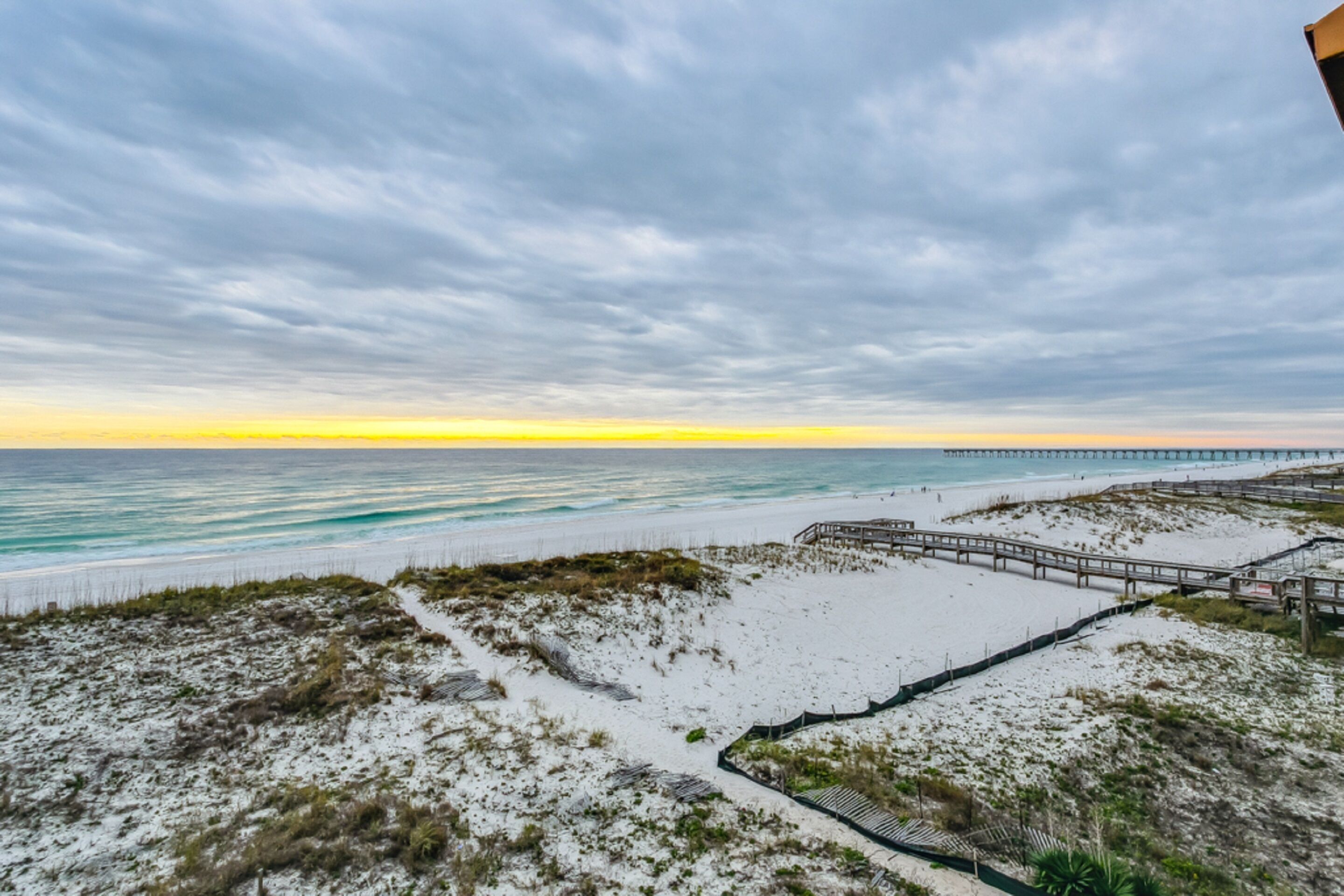 A beach with a wooden boardwalk, sandy dunes, and a pier in the distance.