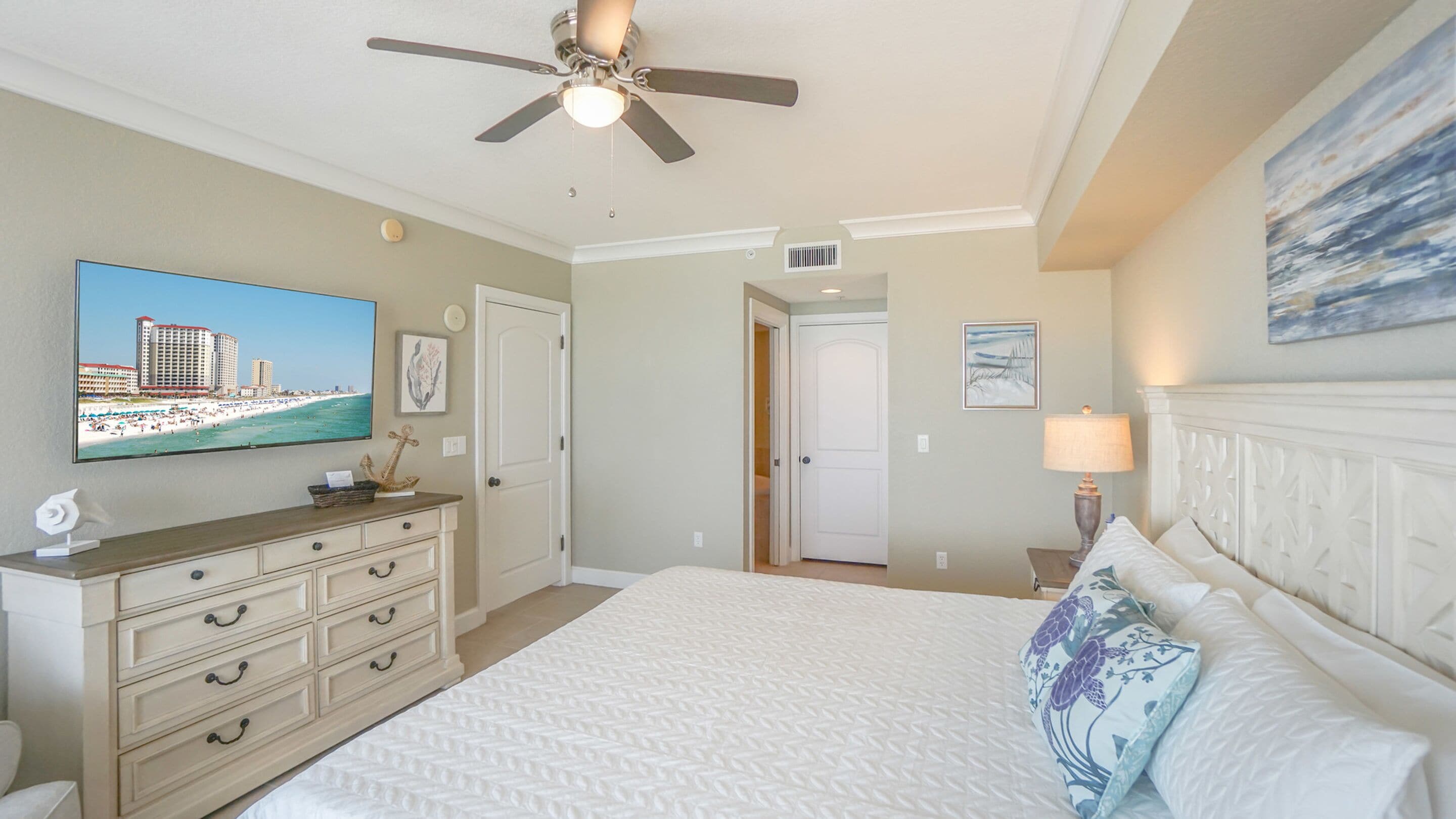 A hotel room with a bed, a dresser, a television displaying a beach scene, a ceiling fan, and a painting on the wall.