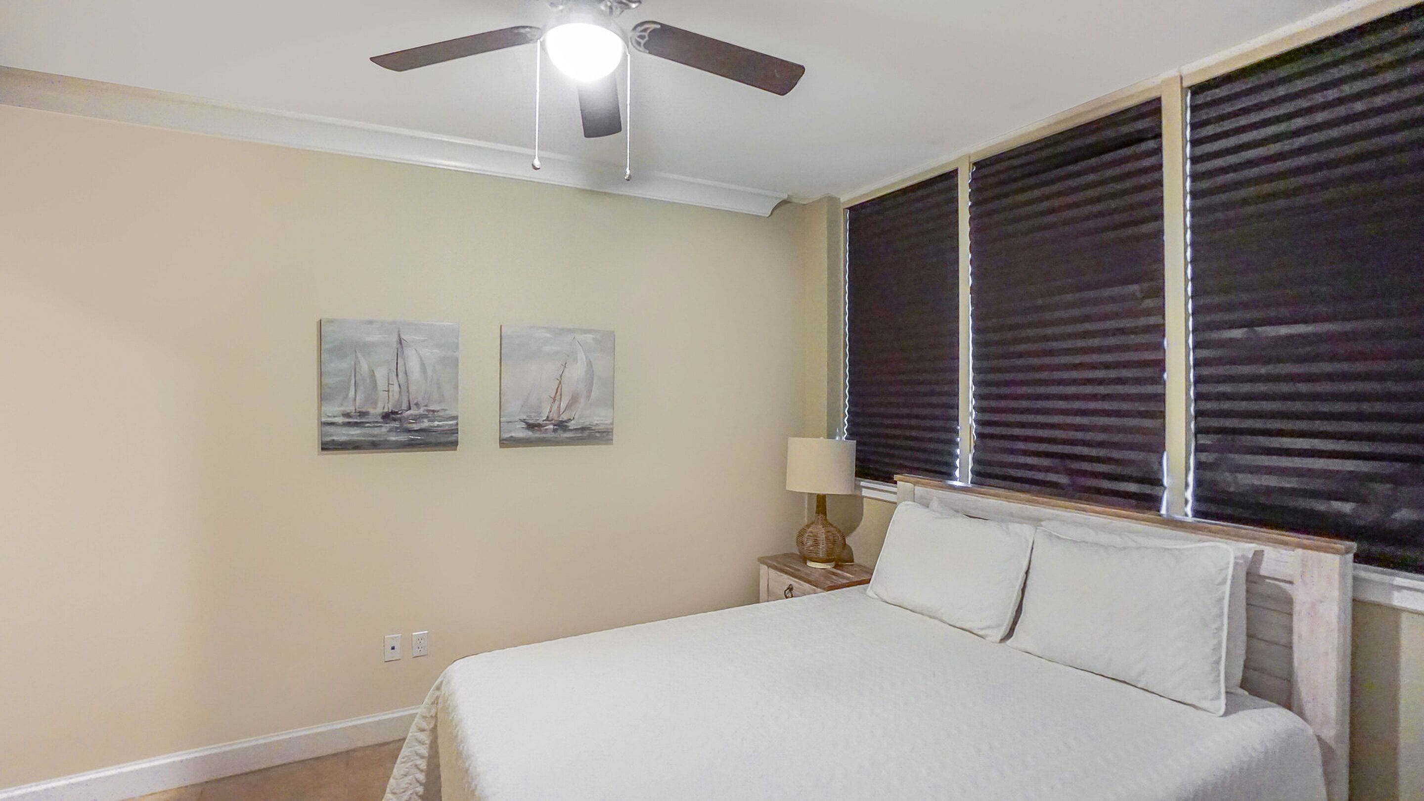 A bedroom with a bed, a ceiling fan, two paintings, a nightstand with a lamp, and a window with blinds.