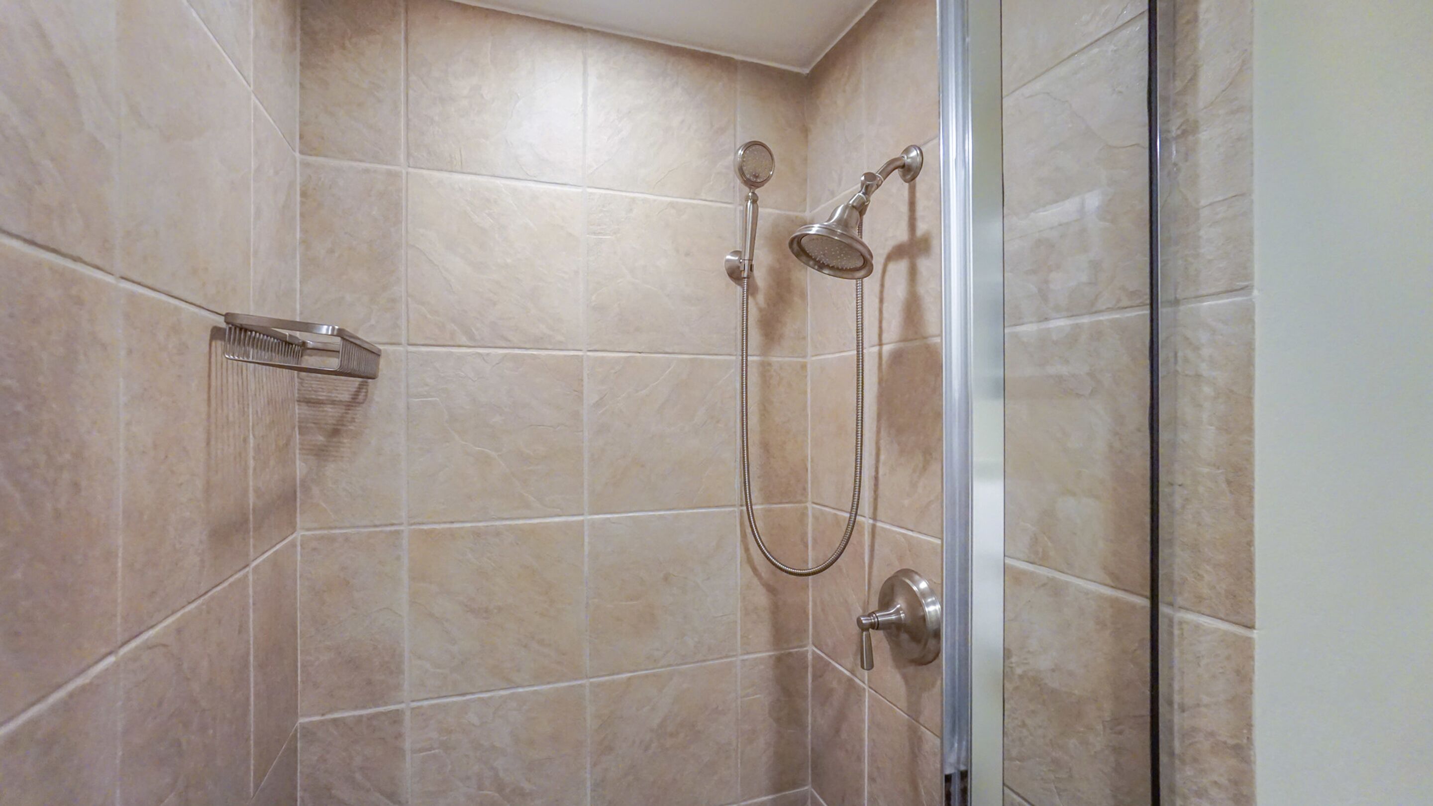Dusche mit Handbrause und fest installierter Duschköpfe, beides an einer Wand mit beigen Fliesen angebracht.