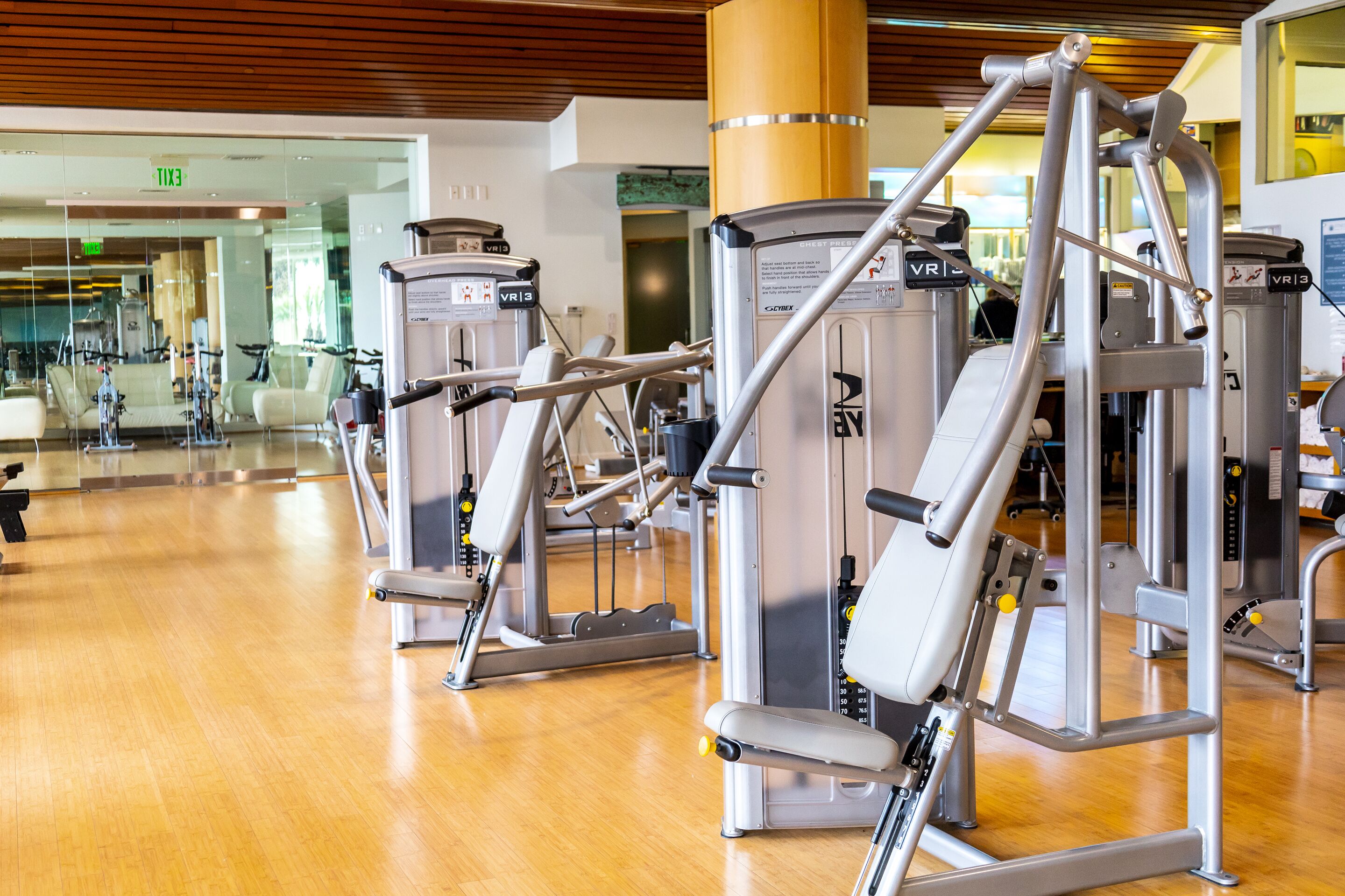 A gym with various exercise machines, wooden flooring, and a visible exit sign.