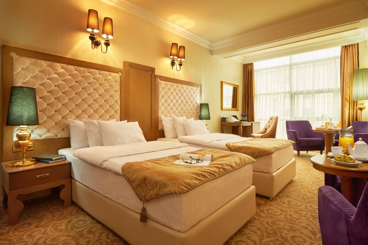 Hotelkamer met een groot bed, twee nachtkastjes, een stoel, een klein tafeltje met een lamp en uitzicht op het buitenleven door grote ramen.