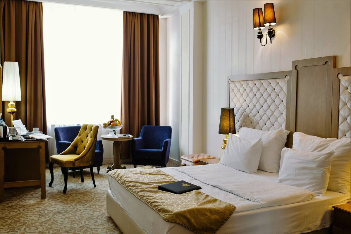 Hotelkamer met een groot bed, een bureau met lamp, een blauwe fauteuil en een kleine ronde tafel met lamp.