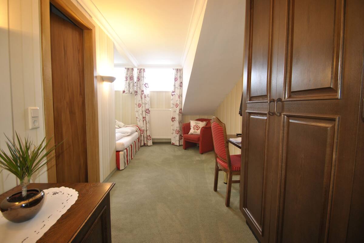 A hallway with a wooden door, a small table with a plant, a red chair, and a bed with a white headboard.