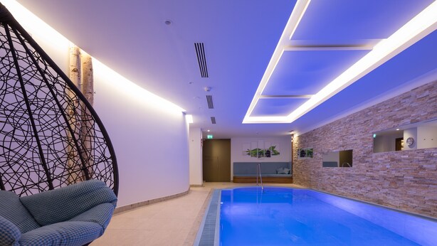 A modern indoor swimming pool with a hanging chair, a stone wall, and recessed lighting. - Hotel Albona Nova (Lech am Arlberg)