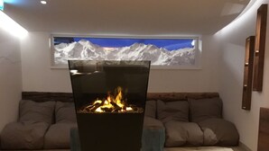 A modern living room with a central fireplace, a cozy sofa set, and a large window with a mountain view. - Hotel Albona Nova (Lech am Arlberg)
