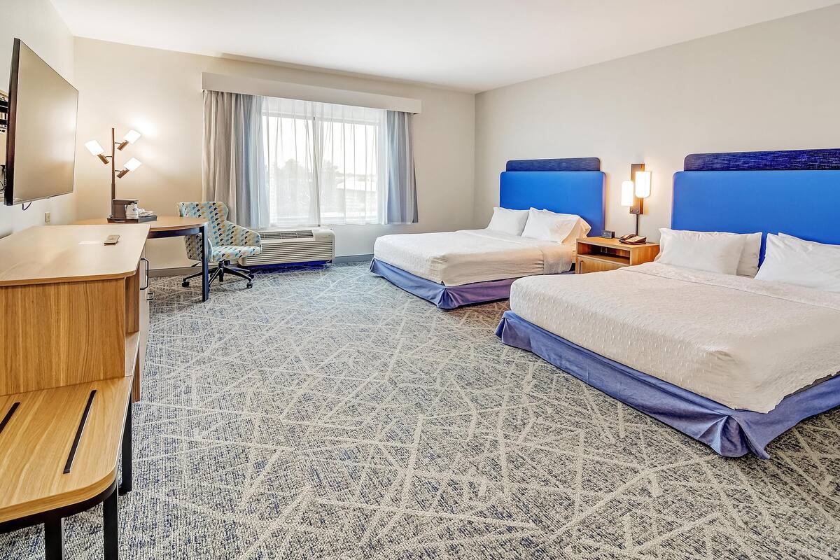 A hotel room with two beds, a desk, a chair, a TV, and a large window with curtains.
