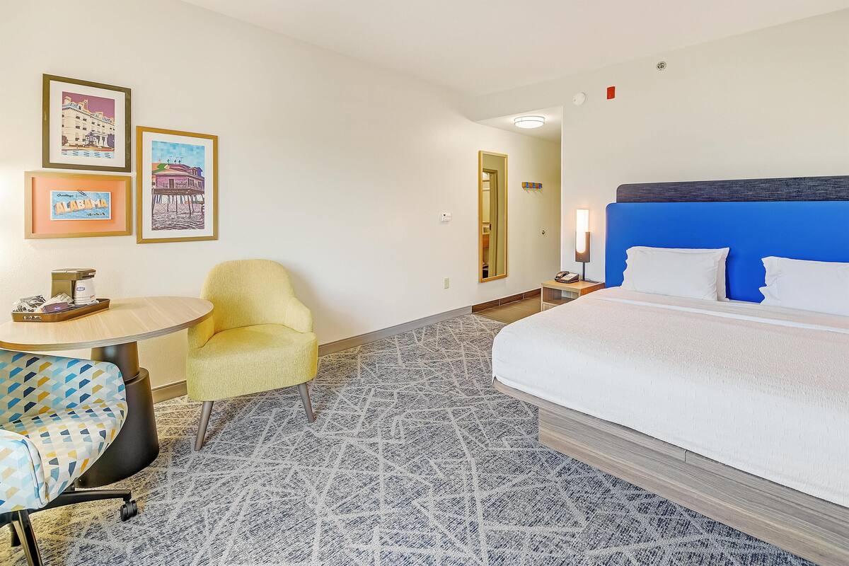 A hotel room with a bed, a chair, a table, and framed pictures on the wall.