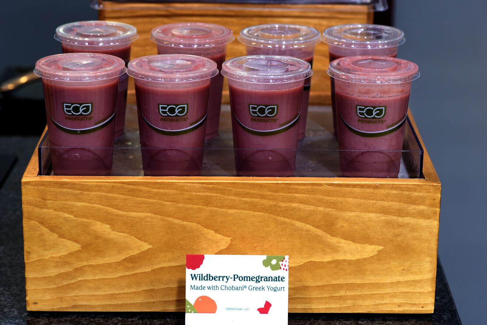 Eine Holzkiste mit sechs Granatapfel-Smoothies in durchsichtigen Plastikbechern mit Deckeln, beschriftet mit 'ECO PRODUCTS'.