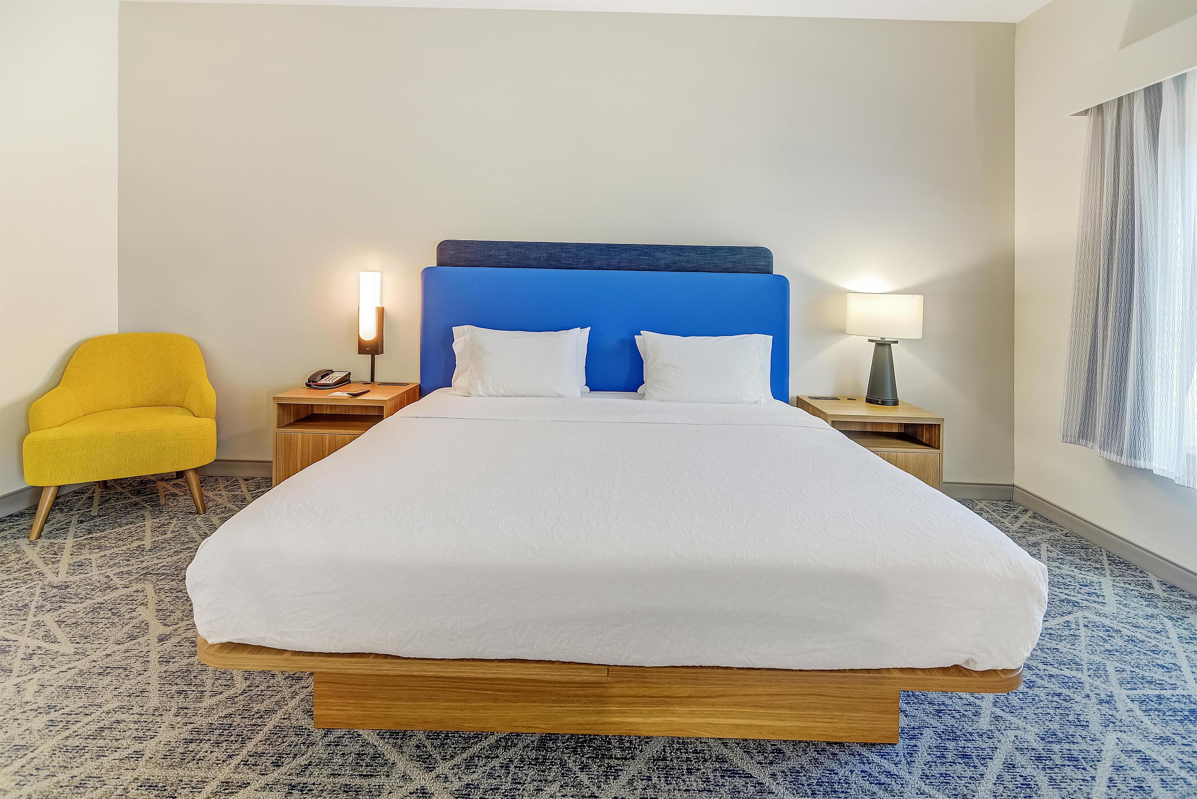 A hotel room with a large bed, two bedside tables, a yellow armchair, and a lamp.