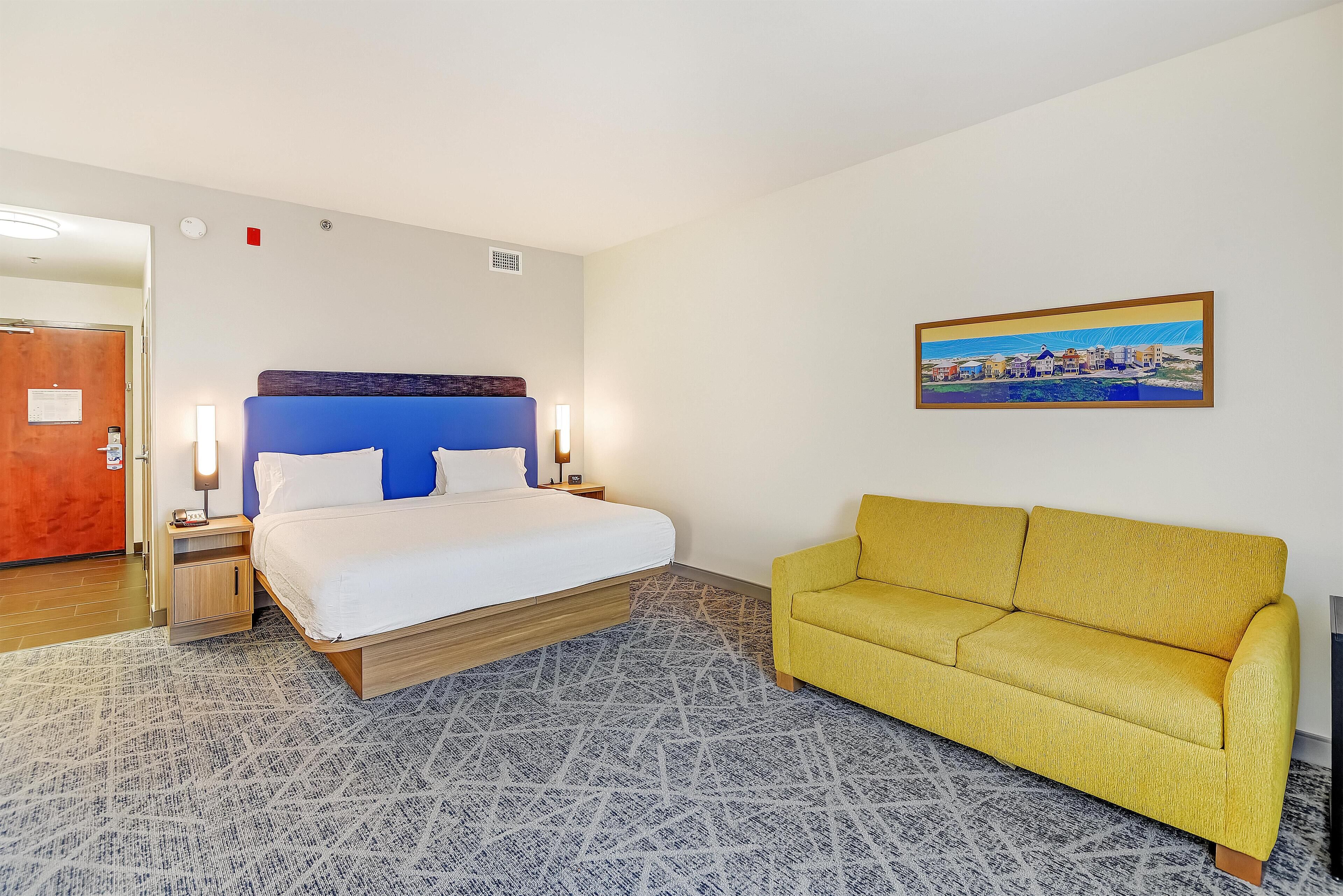 A hotel room with a bed, a yellow sofa, a patterned rug, and a painting on the wall.