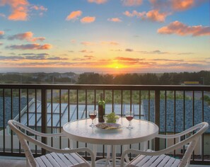 A balcony with a table set for two, overlooking a body of water and a sunset.