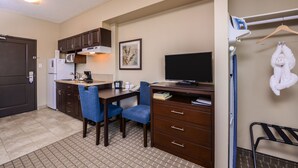 A compact hotel room with a kitchenette, a desk with a chair, a flat-screen TV, and a hanging towel rack. - Chateau All Suites Hotel (Kindersley)