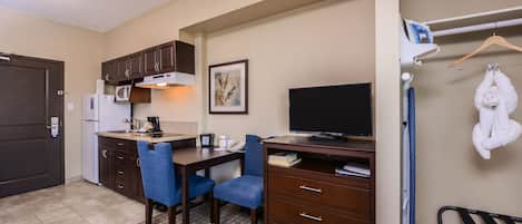 Une chambre d’hôtel compacte comprenant un coin cuisine, un bureau avec une chaise, un téléviseur à écran plat et un porte-serviettes mural.