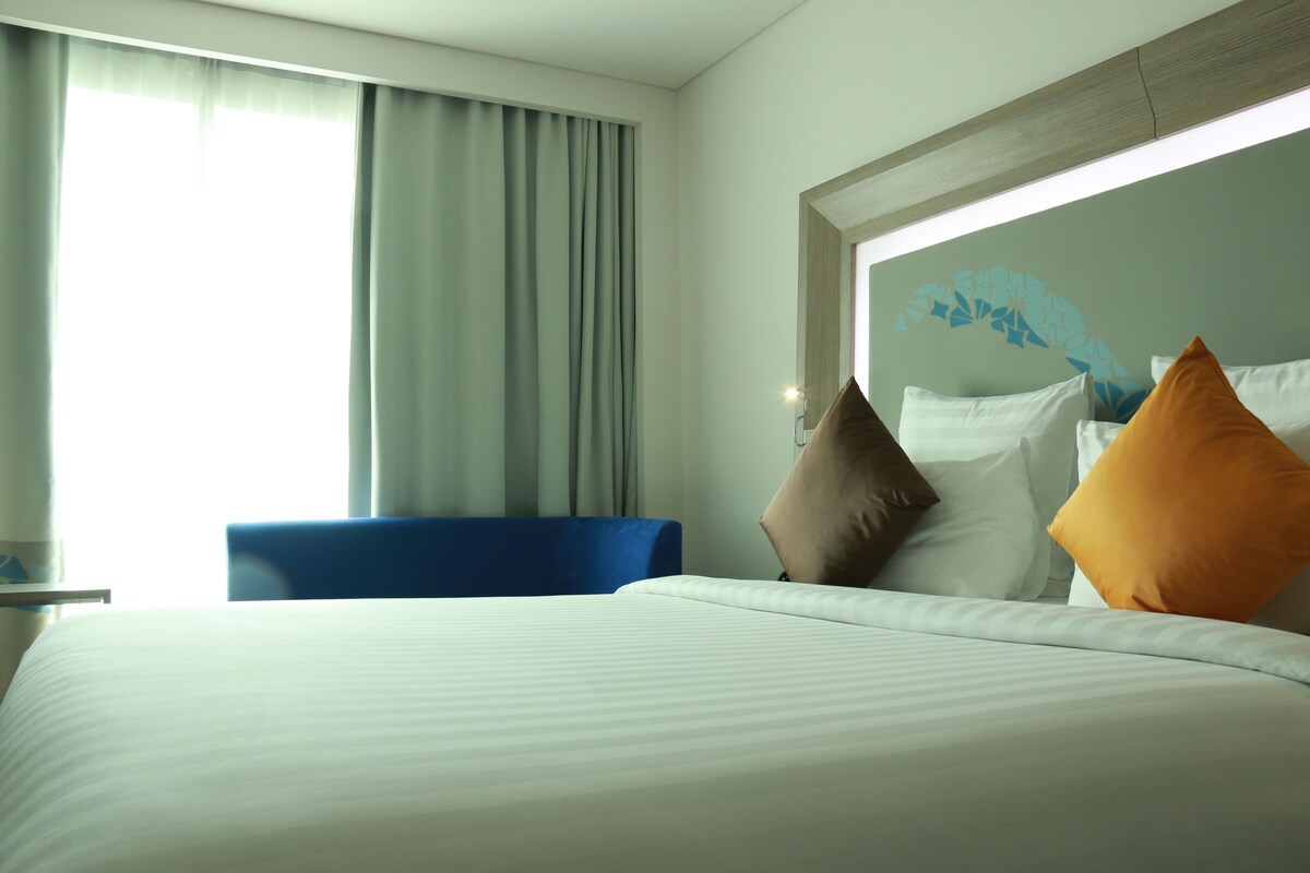 A hotel room with a bed, a blue chair, and a large window with curtains.