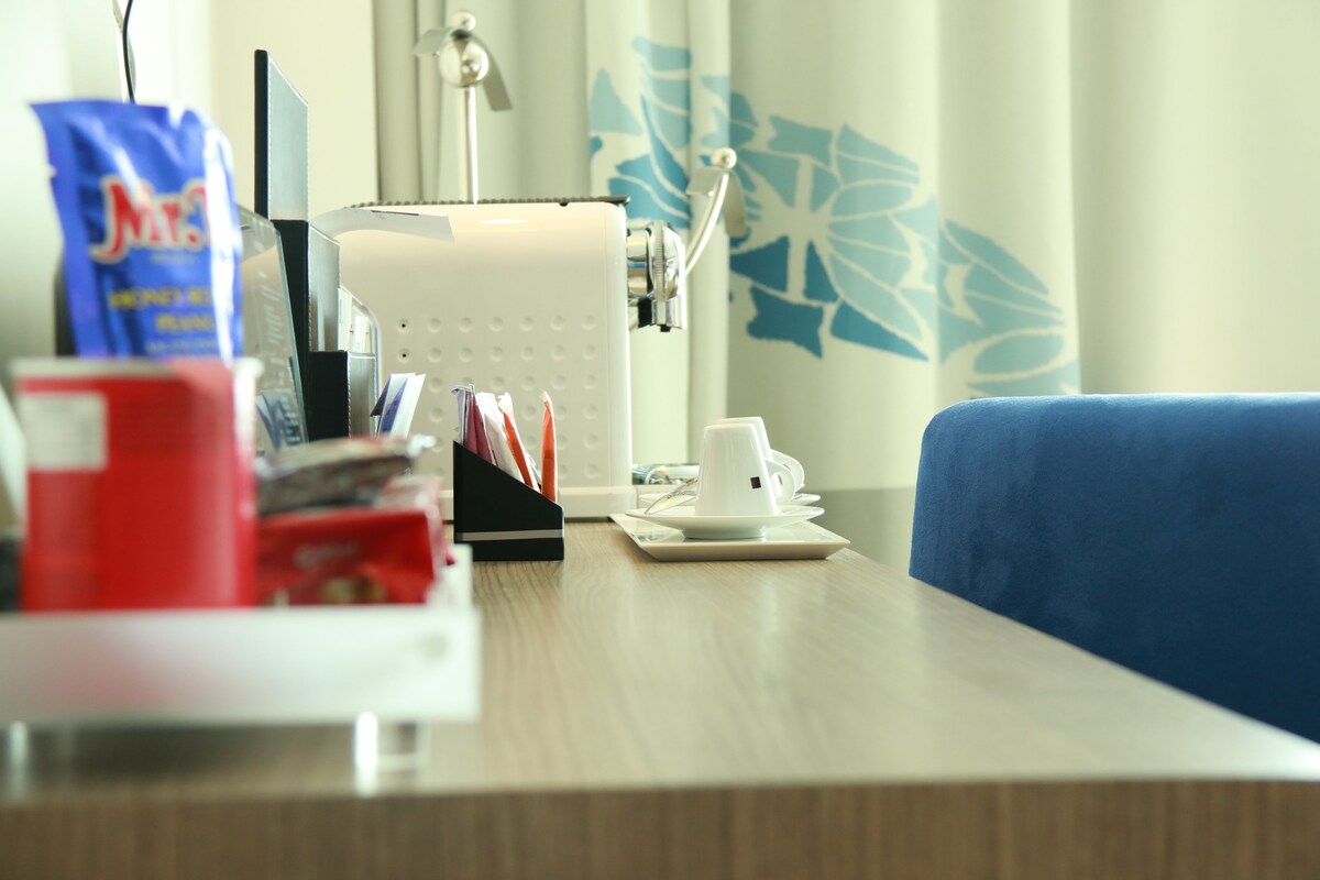 A hotel room with a wooden table, a white coffee maker, a red cup, and a blue couch.