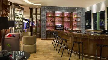 Un'area bar moderna con un bancone curvo, sgabelli da bar e un televisore. Sono presenti scaffali con bottiglie e una parete decorativa con un motivo geometrico.
