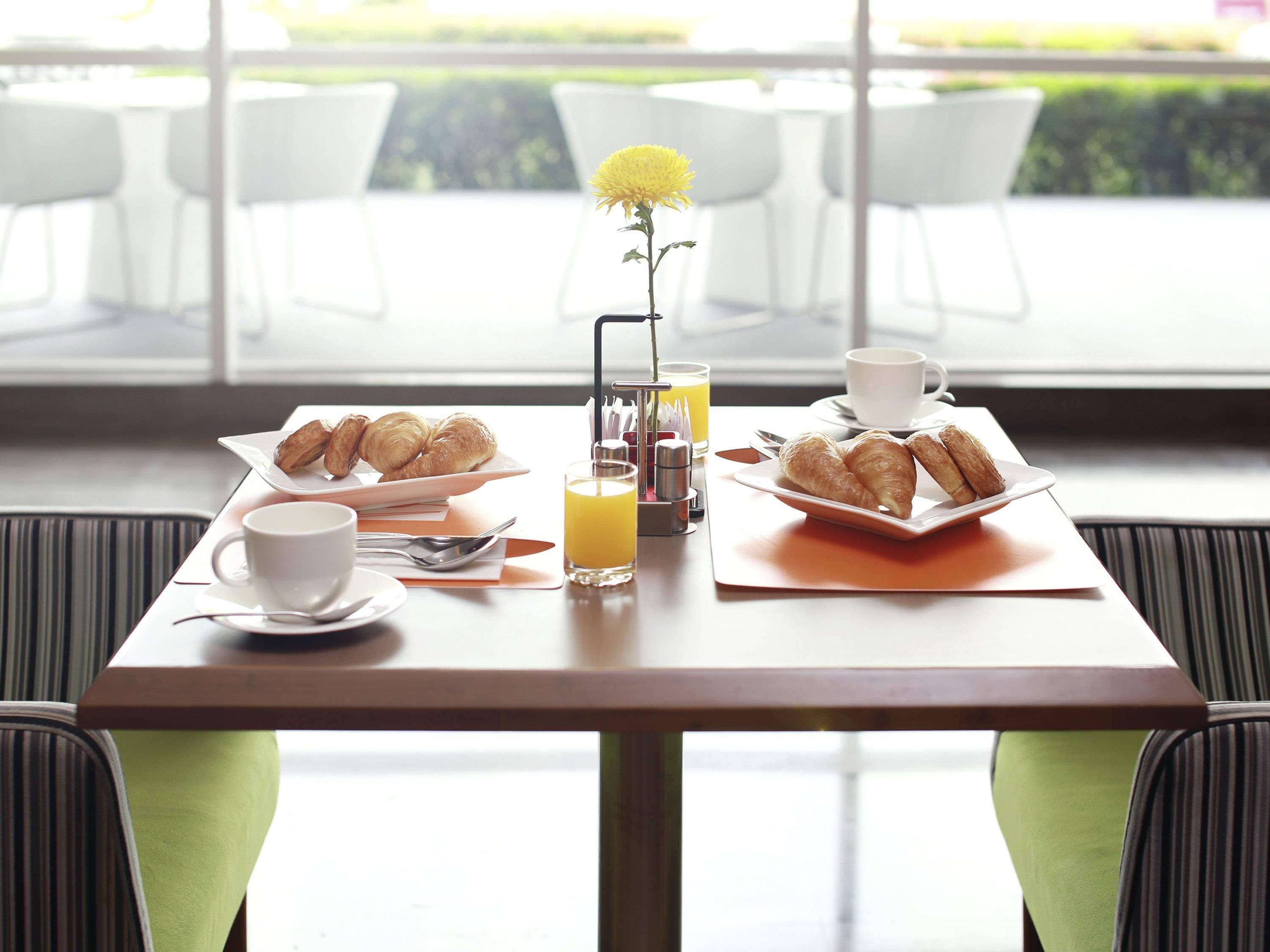 Una mesa de desayuno con cruasanes, una taza de café, un vaso de jugo de naranja y un centro de mesa floral.