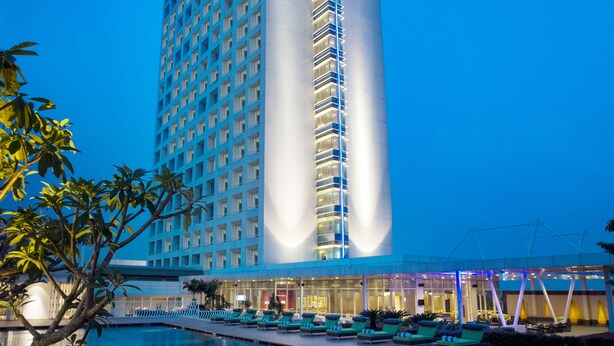 Hotel alto com área de piscina, apresentando design moderno e uma faixa vertical de luz marcante no edifício.